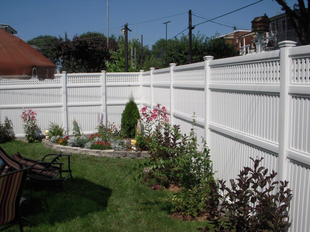 Inter Clôtures Sentinelle A backyard with a white vinyl privacy fence, showcasing expert fencing solutions, landscaped garden beds along the fence, and outdoor chairs on a green lawn. Fence Installation Quebec