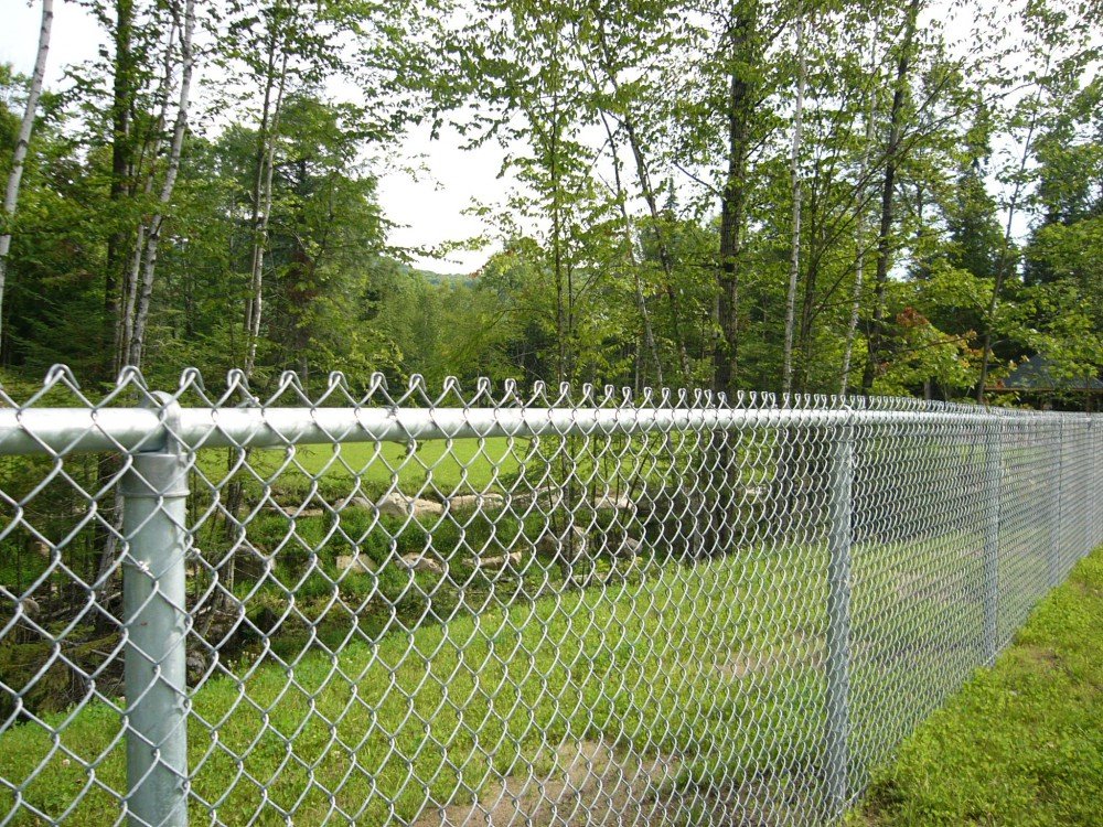 Inter Clôtures Sentinelle A chain-link fence, ideal for residential fencing, extends across a grassy area with trees in the background. Fence Installation Quebec