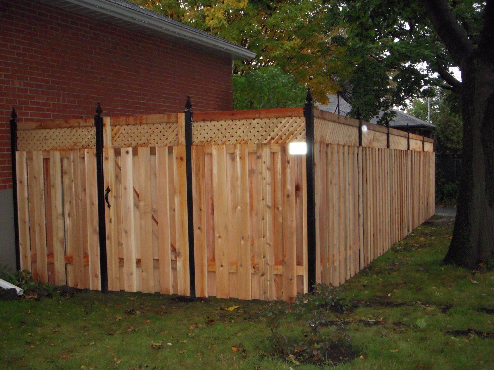 Inter Clôtures Sentinelle A newly built wooden privacy fence with lattice panels on top surrounds the side yard of a brick house, bordered by grass and trees, showcasing quality fencing solutions for added security and style. Fence Installation Quebec
