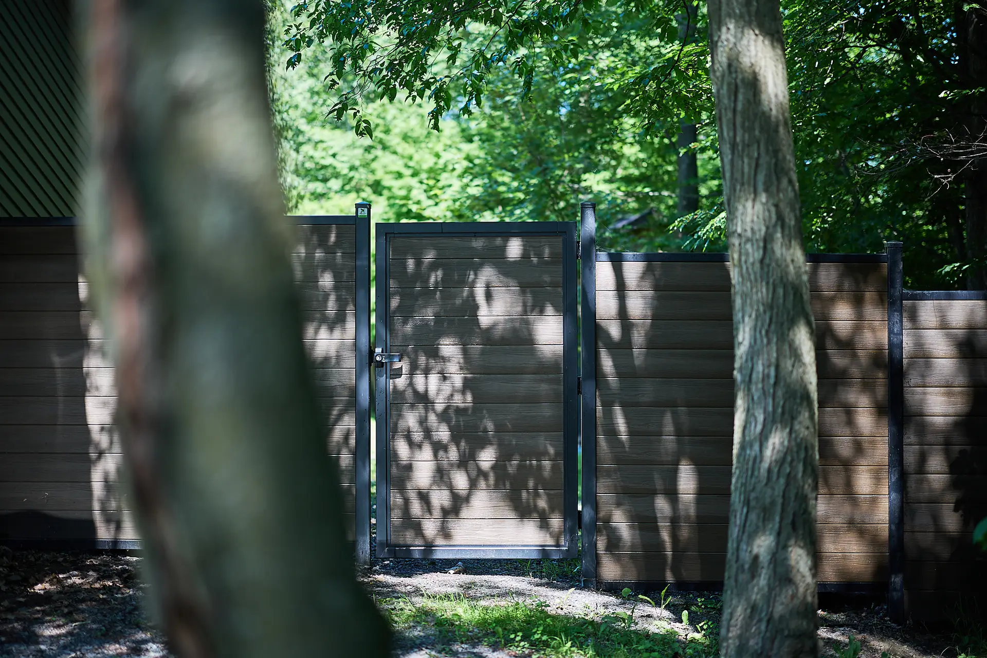 Inter Clôtures Sentinelle A closed wooden gate set in a tall wooden fence is surrounded by trees, with sunlight casting shadows on the surfaces. Fence Installation Quebec