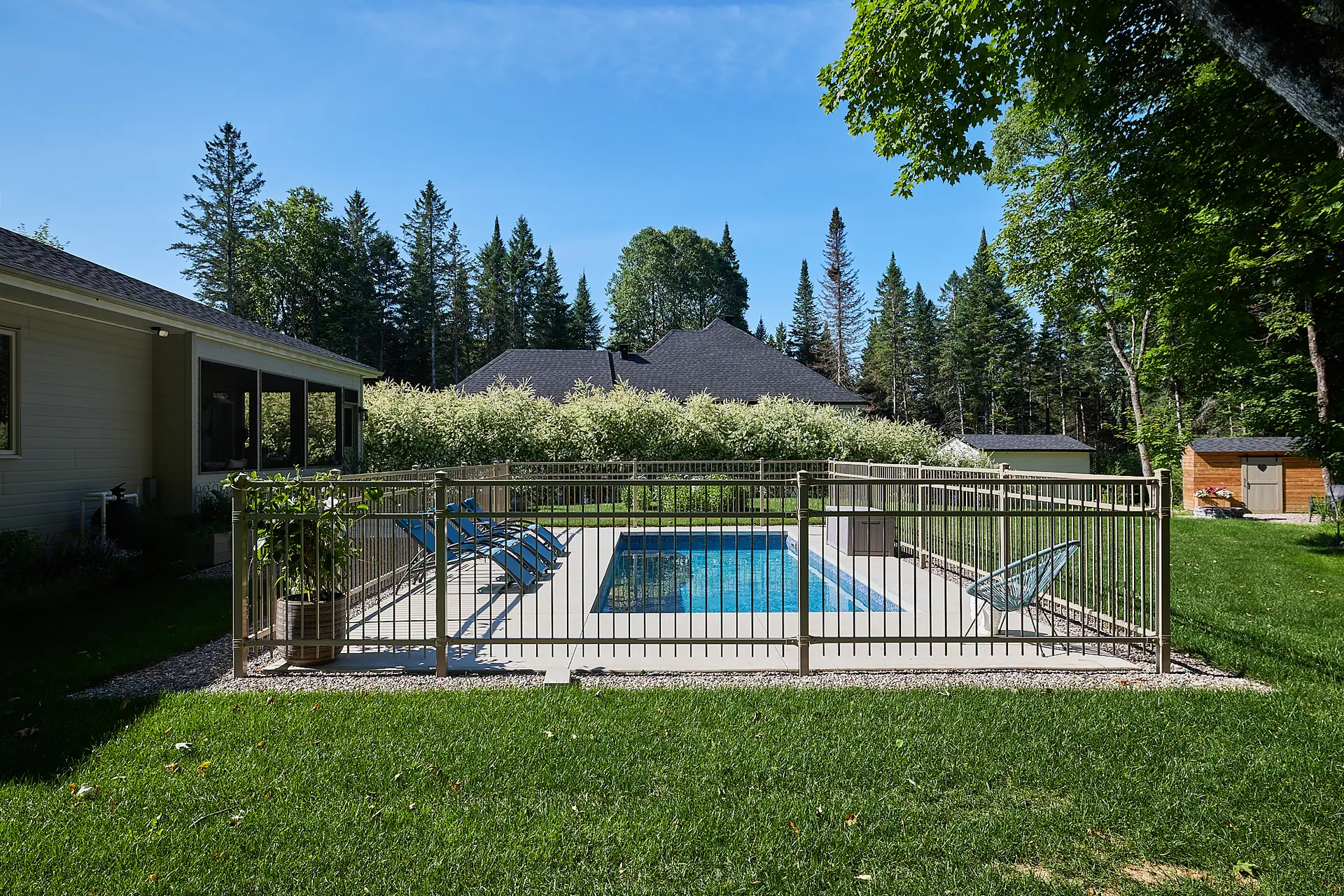 Inter Clôtures Sentinelle A fenced backyard swimming pool with lounge chairs, surrounded by grass, trees, and houses under a clear blue sky. Fence Installation Quebec