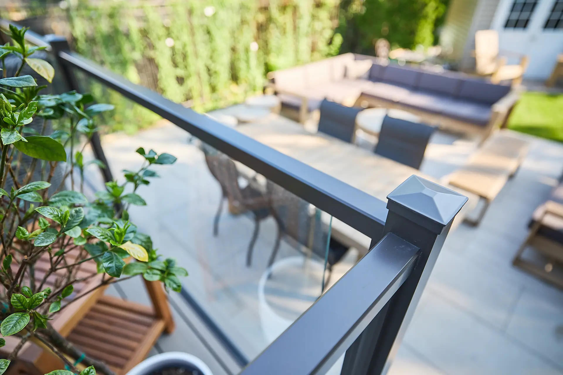 Inter Clôtures Sentinelle Close-up of a metal and glass balcony railing with patio furniture, potted plants, and green foliage visible in the background. Fence Installation Quebec