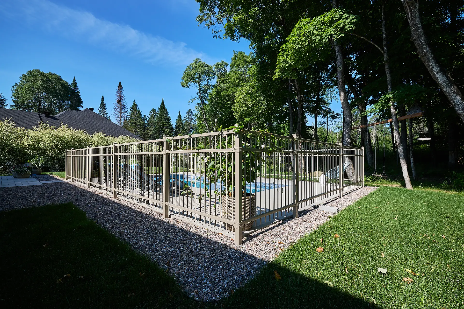 Inter Clôtures Sentinelle A fenced-in outdoor swimming pool is surrounded by lawn, gravel paths, and trees under a clear blue sky. Fence Installation Quebec