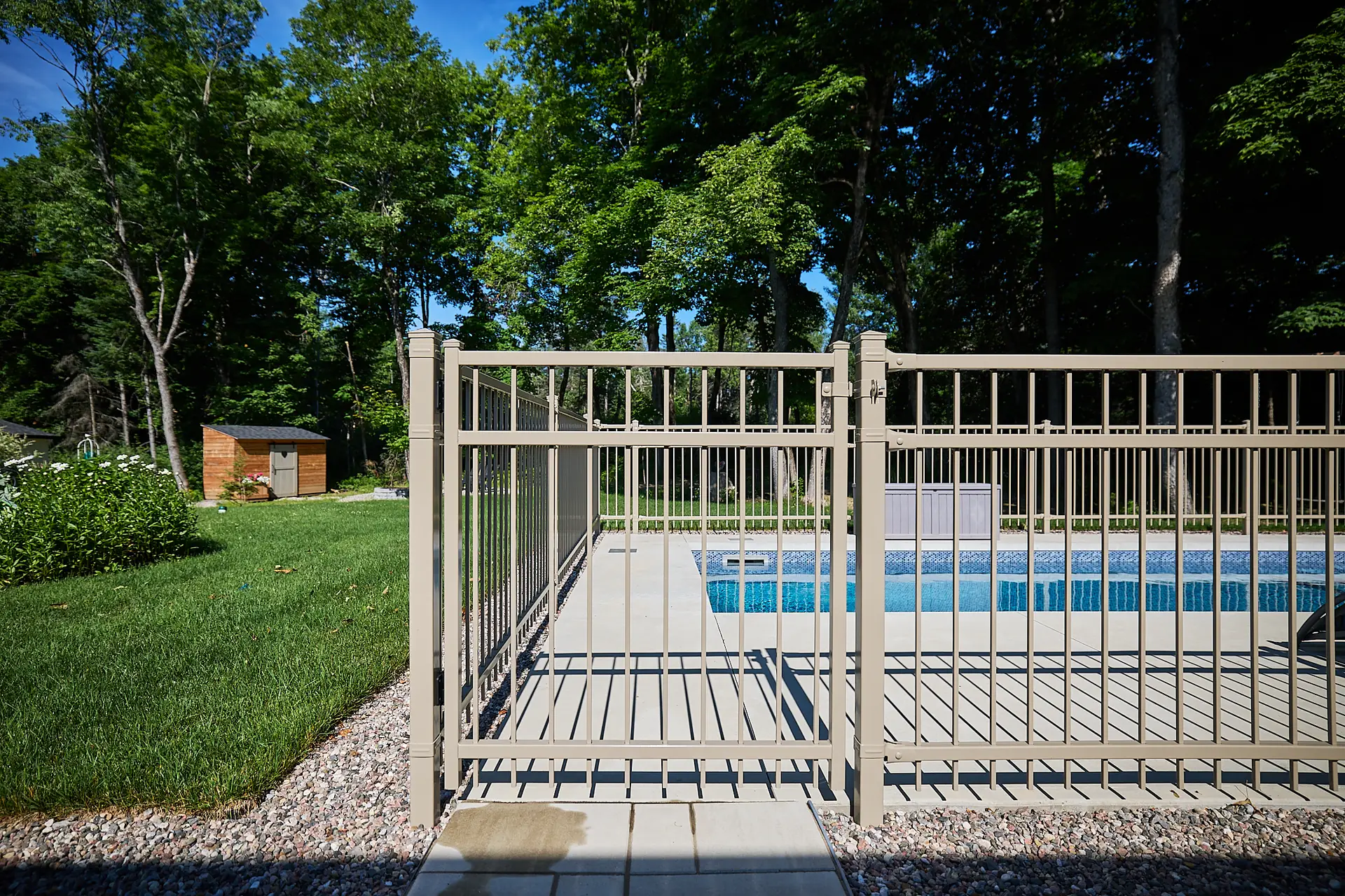 Inter Clôtures Sentinelle Metal fence gate surrounding an outdoor swimming pool with green grass and trees in the background on a sunny day. Fence Installation Quebec