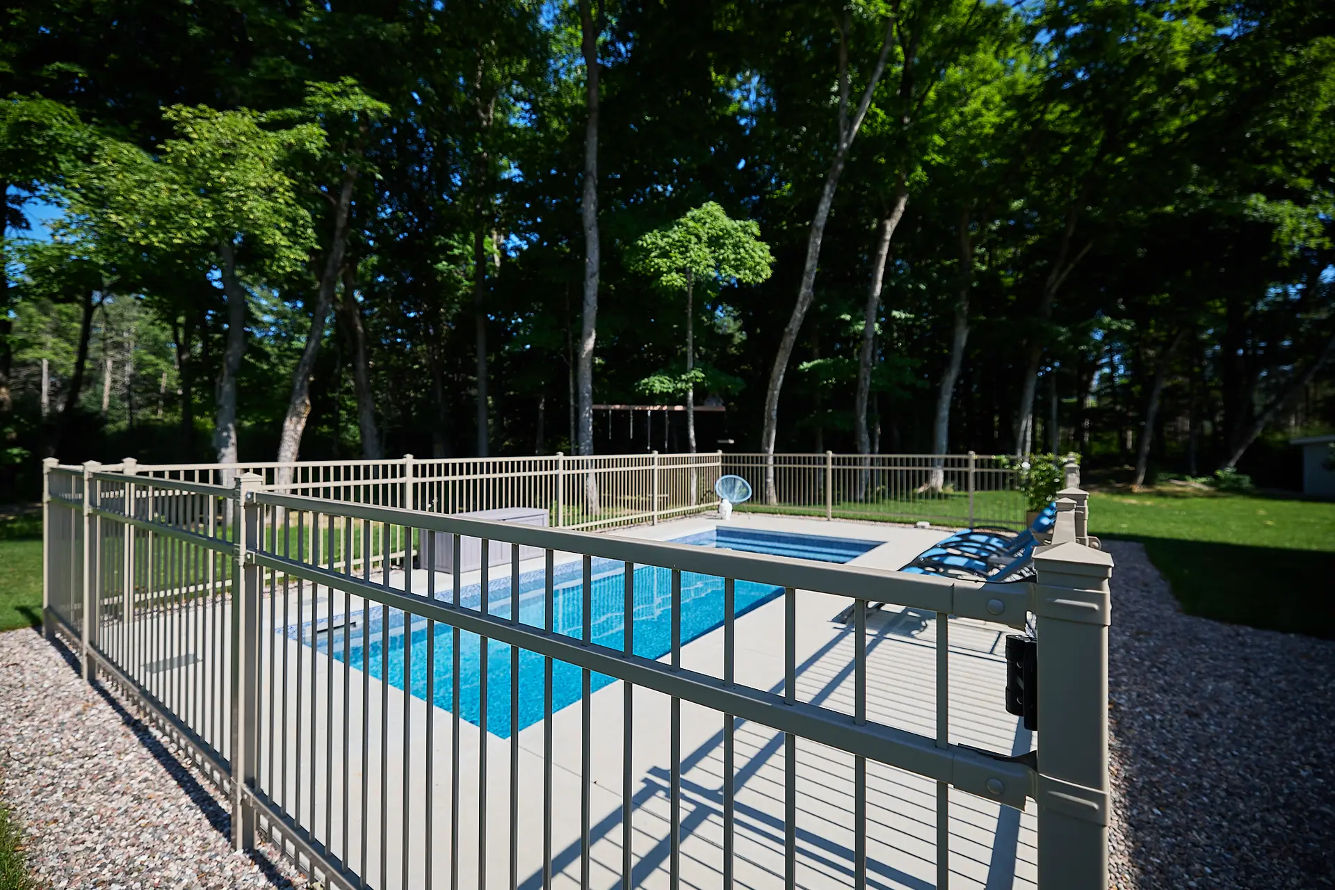 Inter Clôtures Sentinelle A small rectangular swimming pool surrounded by a metal safety fence, with two lounge chairs nearby, set in a backyard with trees in the background. Fence Installation Quebec