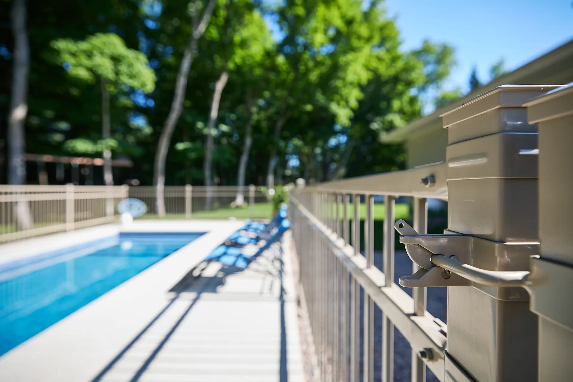 Inter Clôtures Sentinelle Close-up of a locked pool gate latch with a view of a fenced-in outdoor swimming pool and lounge chairs in the background surrounded by trees. Fence Installation Quebec