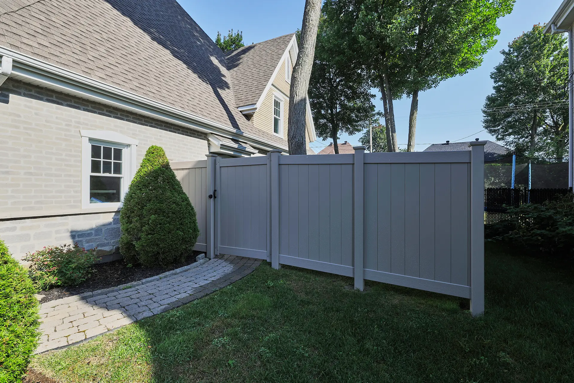 Inter Clôtures Sentinelle Gray privacy fence with a gate separates a side yard area next to a house; pavers form a walkway beside a trimmed bush and green lawn. Fence Installation Quebec
