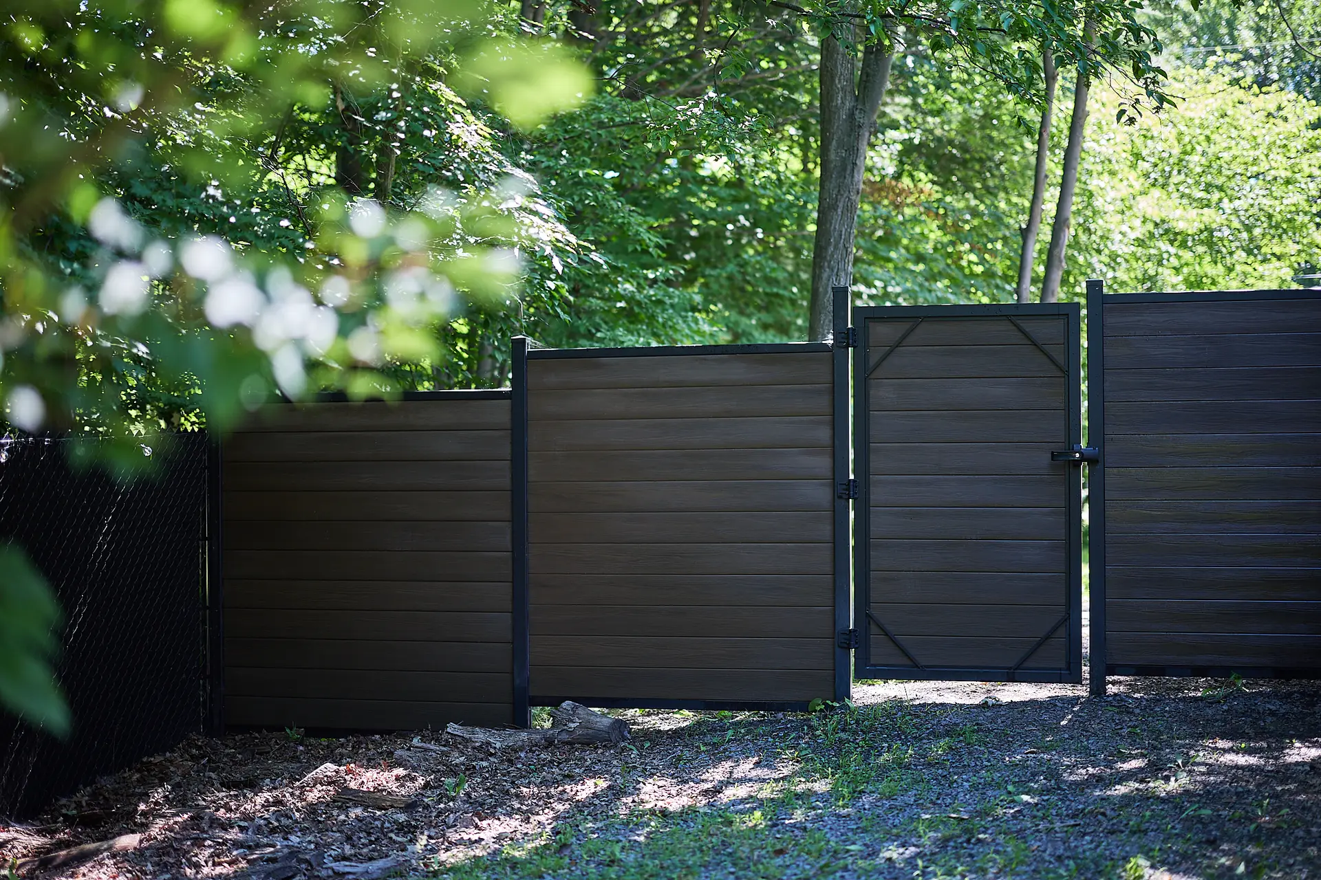 Inter Clôtures Sentinelle Brown privacy fence with a closed gate stands in a shaded, wooded outdoor area with sunlight filtering through green trees. Fence Installation Quebec