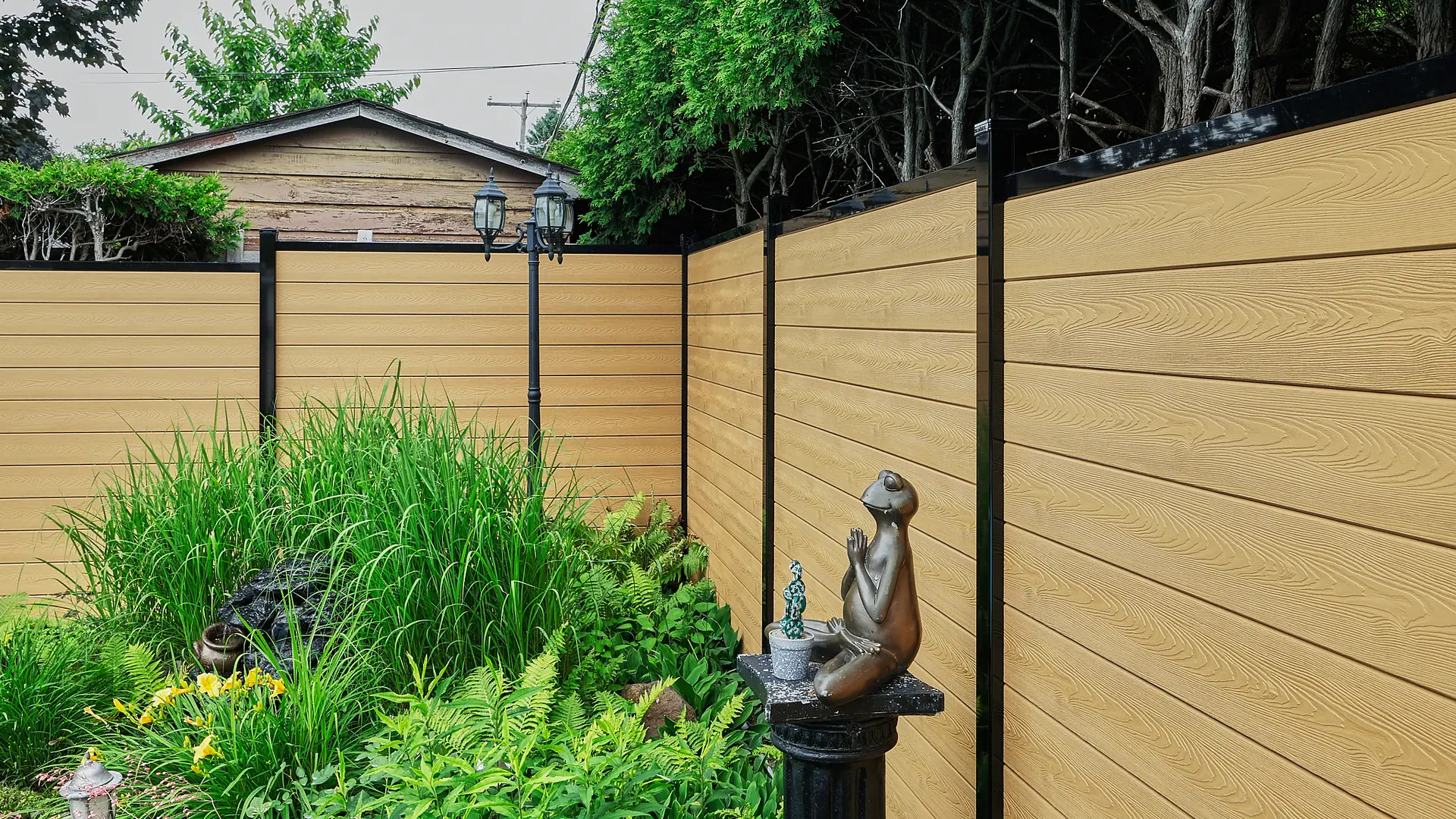 Inter Clôtures Sentinelle A small backyard garden with tall grass, a bronze frog statue on a pedestal, a lamp post, and tan horizontal panel fencing. Fence Installation Quebec