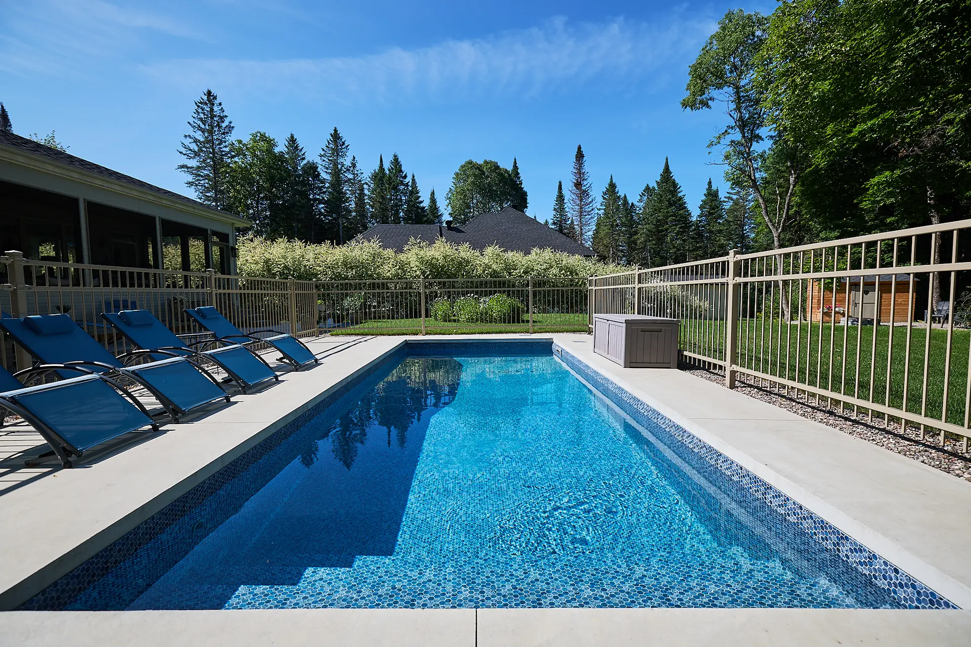 Inter Clôtures Sentinelle Rectangular outdoor swimming pool with clear blue water, surrounded by a metal fence, lounge chairs, and greenery in the background under a sunny sky. Fence Installation Quebec