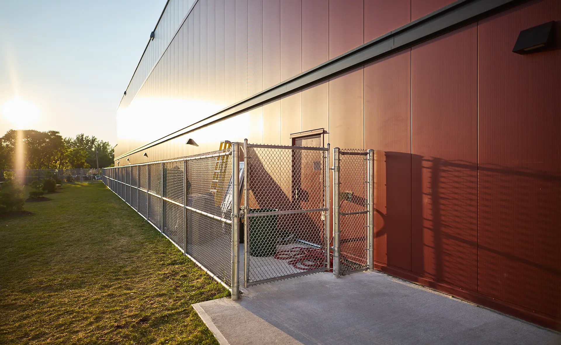 Inter Clôtures Sentinelle A fenced utility area with equipment sits beside a large red industrial building at sunset, with shadows cast on the wall and grass. Fence Installation Quebec