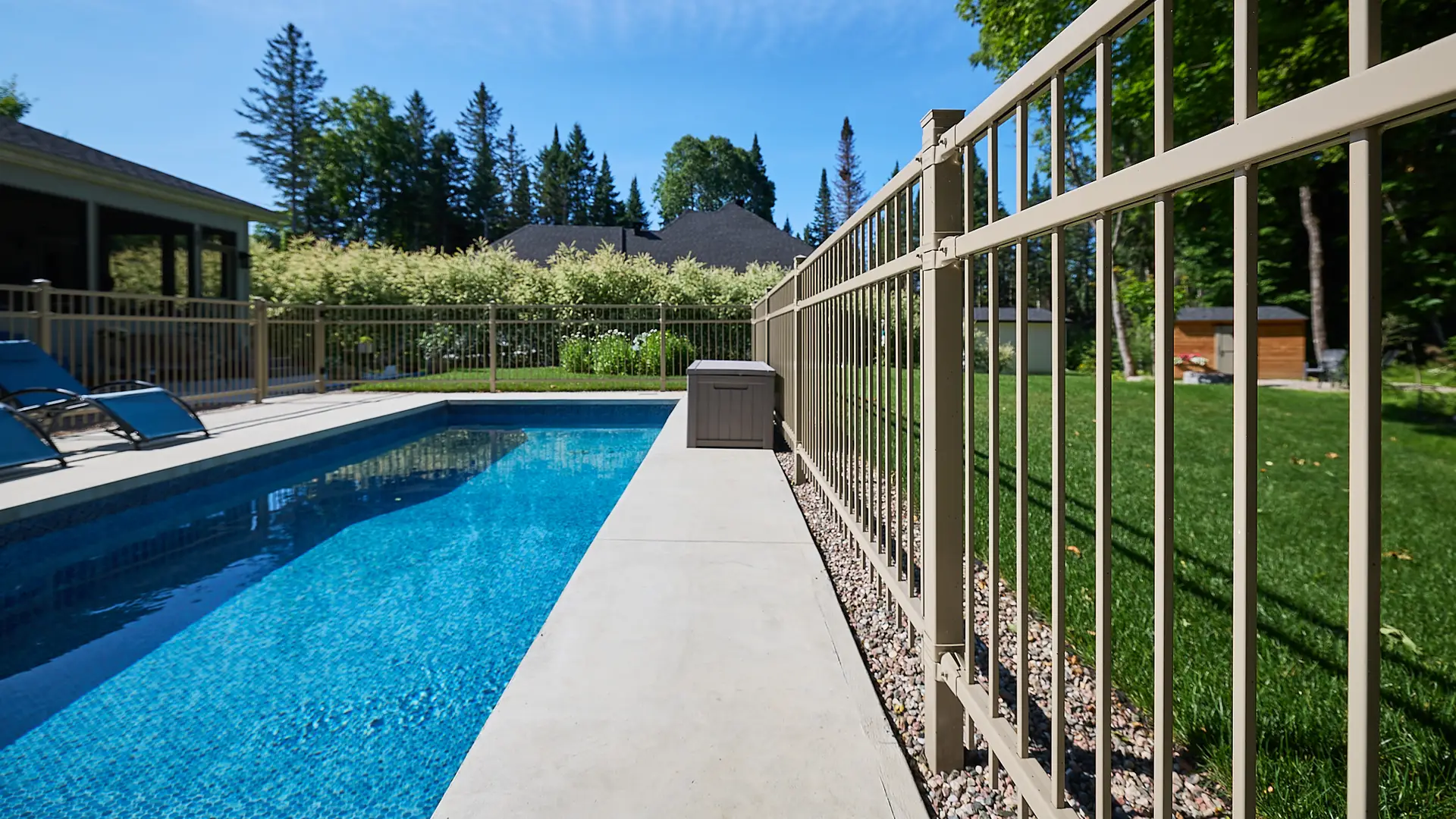 Inter Clôtures Sentinelle A fenced outdoor swimming pool with clear blue water, surrounded by a concrete walkway and green lawn, under a bright sunny sky. Fence Installation Quebec