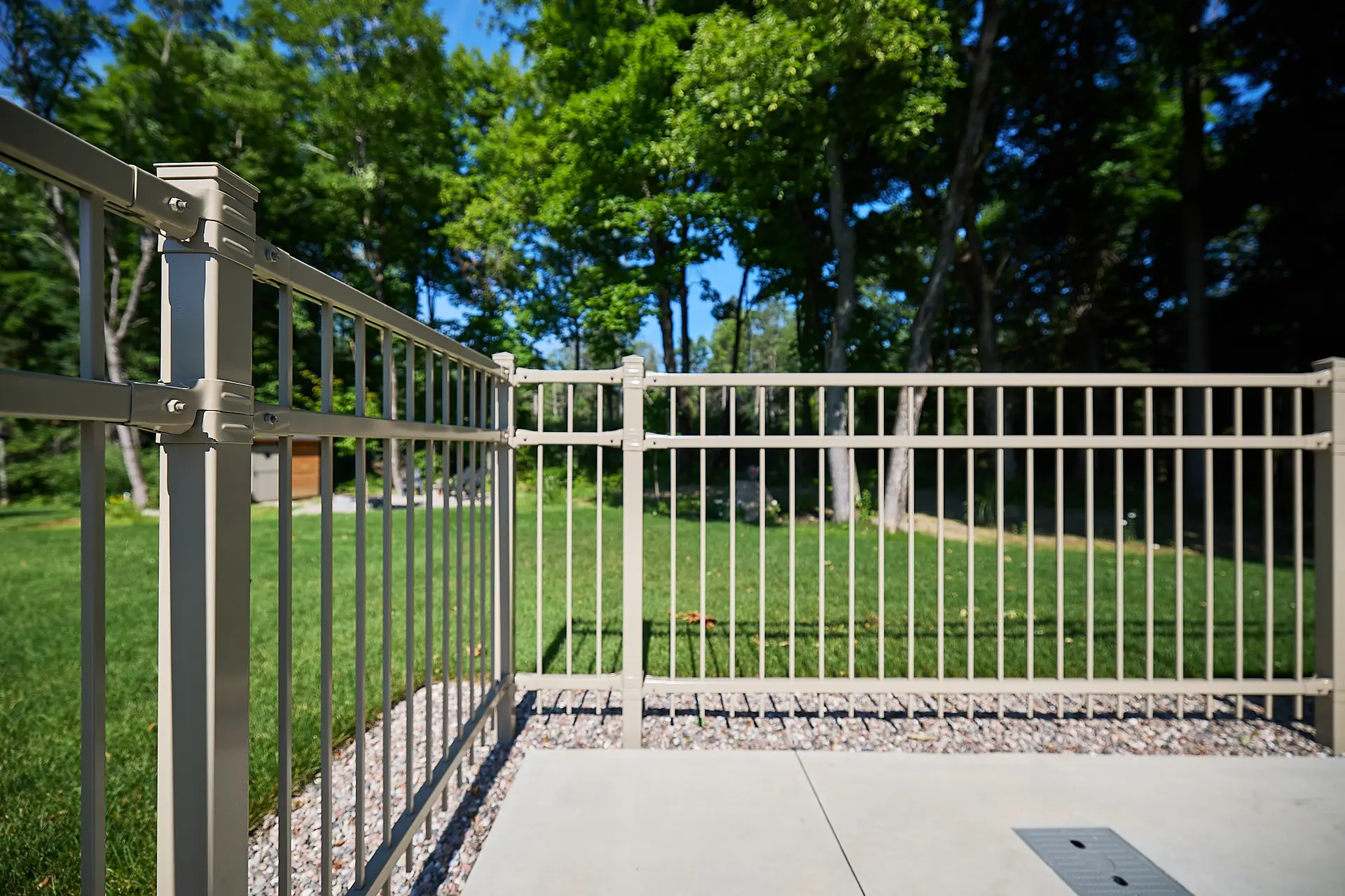 Inter Clôtures Sentinelle Metal fence encloses a small patio area with concrete flooring, surrounded by green grass, trees, and bright sunlight in the background. Fence Installation Quebec