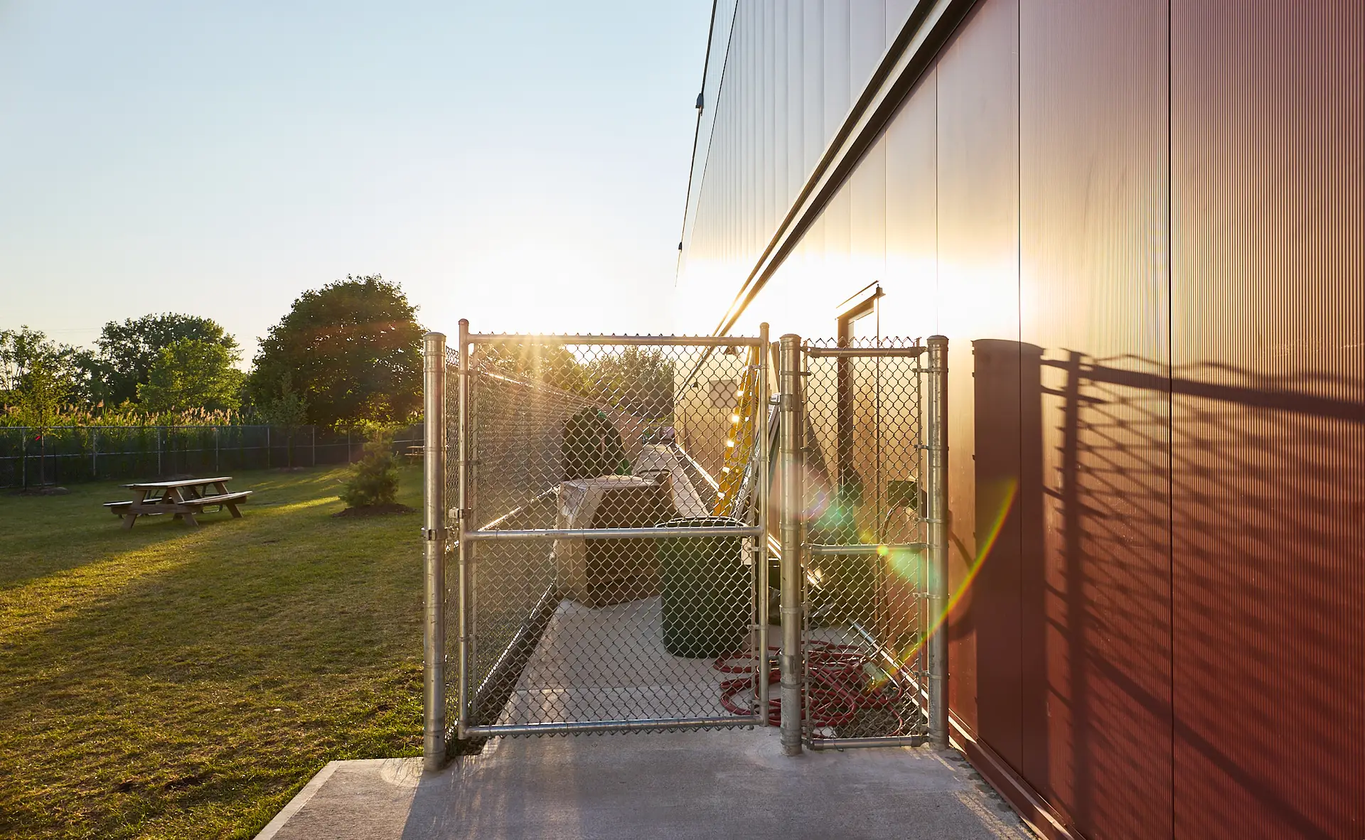 Inter Clôtures Sentinelle Outdoor fenced area next to a red building, with picnic tables and trees in the background, photographed at sunset. Fence Installation Quebec