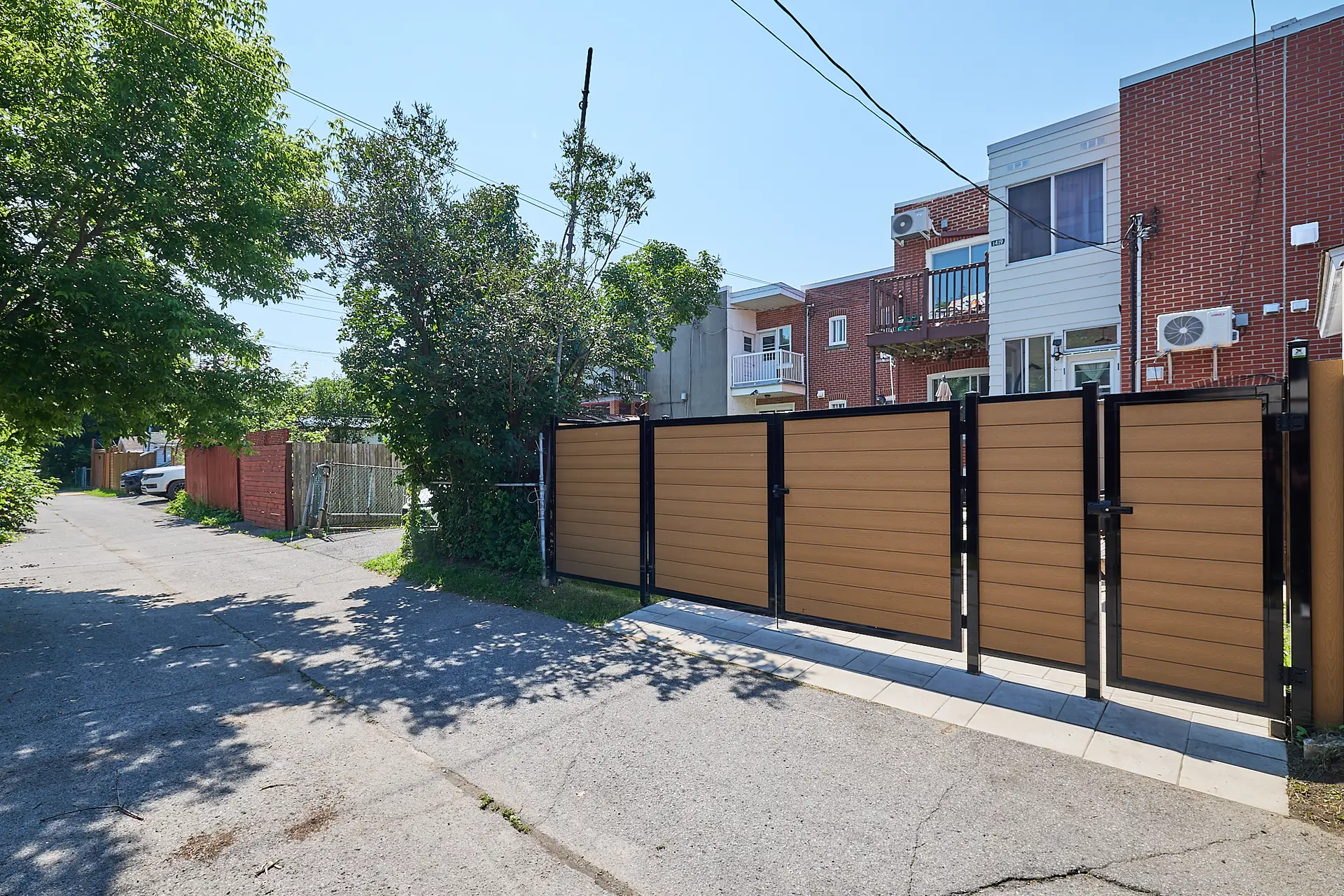 Inter Clôtures Sentinelle A brown and black metal gate blocks a driveway entrance next to a red brick apartment building on a sunny day. Fence Installation Quebec