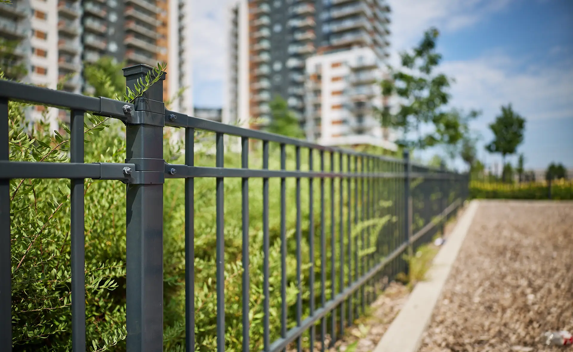 Inter Clôtures Sentinelle Black metal fence with greenery behind it, in front of modern apartment buildings on a sunny day. The image is taken from a low angle, showing the fence running into the distance. Fence Installation Quebec