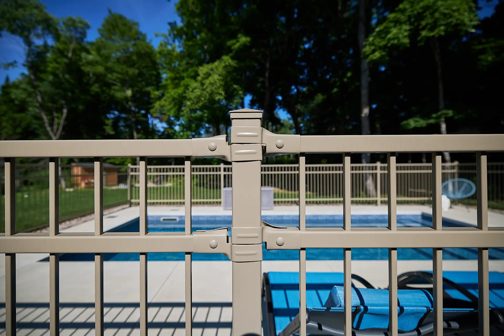 Inter Clôtures Sentinelle A beige metal fence with a gate lock surrounds a backyard swimming pool, with patio chairs and trees visible in the background. Fence Installation Quebec