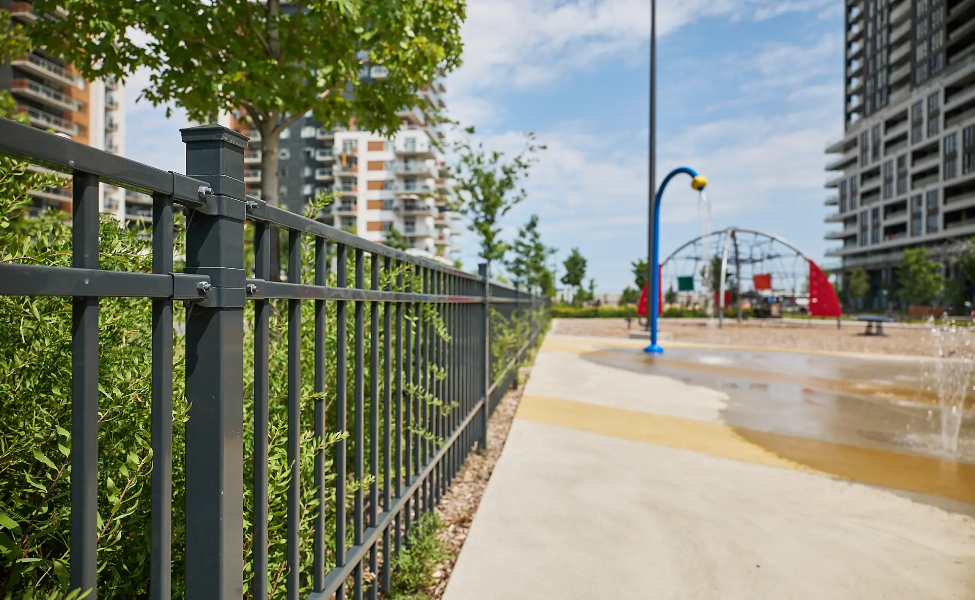 Inter Clôtures Sentinelle Black metal fence beside a concrete path, with nearby greenery, modern apartment buildings, and a splash pad playground in the background. Fence Installation Quebec