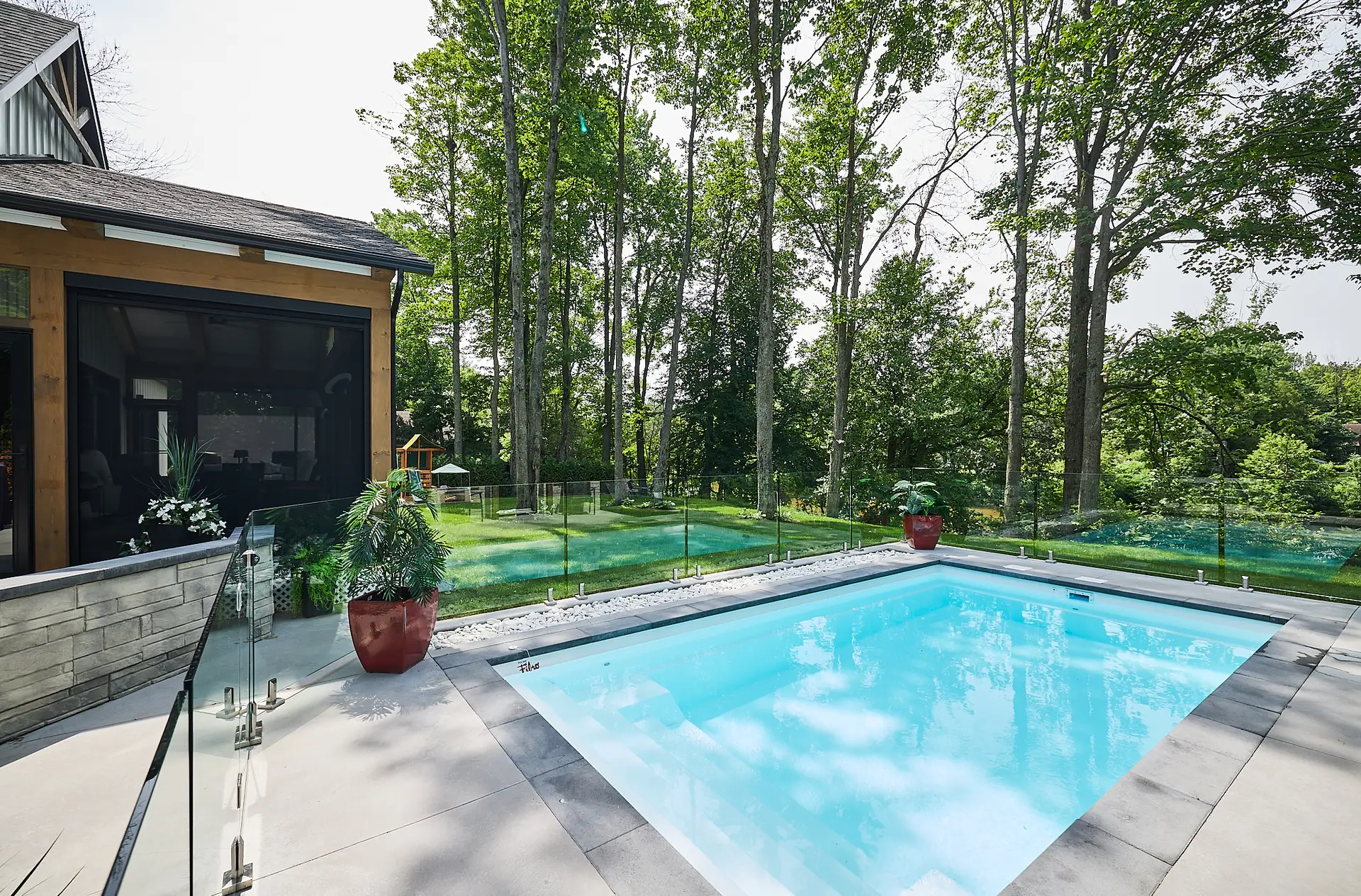 Inter Clôtures Sentinelle Rectangular backyard pool with glass fence, surrounded by trees and lawn, next to a modern house with screened porch. Fence Installation Quebec