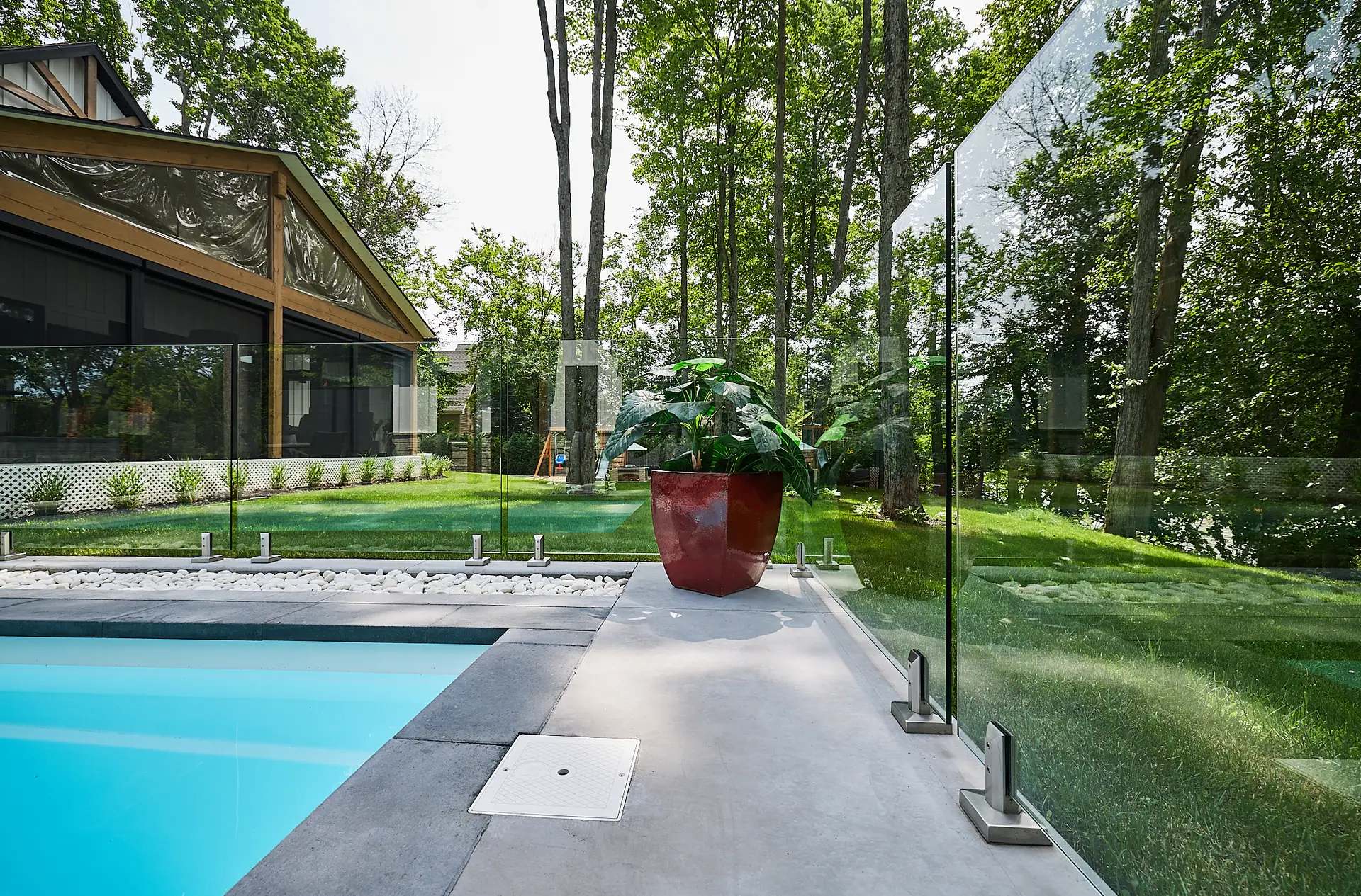 Inter Clôtures Sentinelle A glass fence encloses a modern pool area with a red potted plant beside the water. A house with large windows and a green lawn is visible in the background, surrounded by tall trees. Fence Installation Quebec