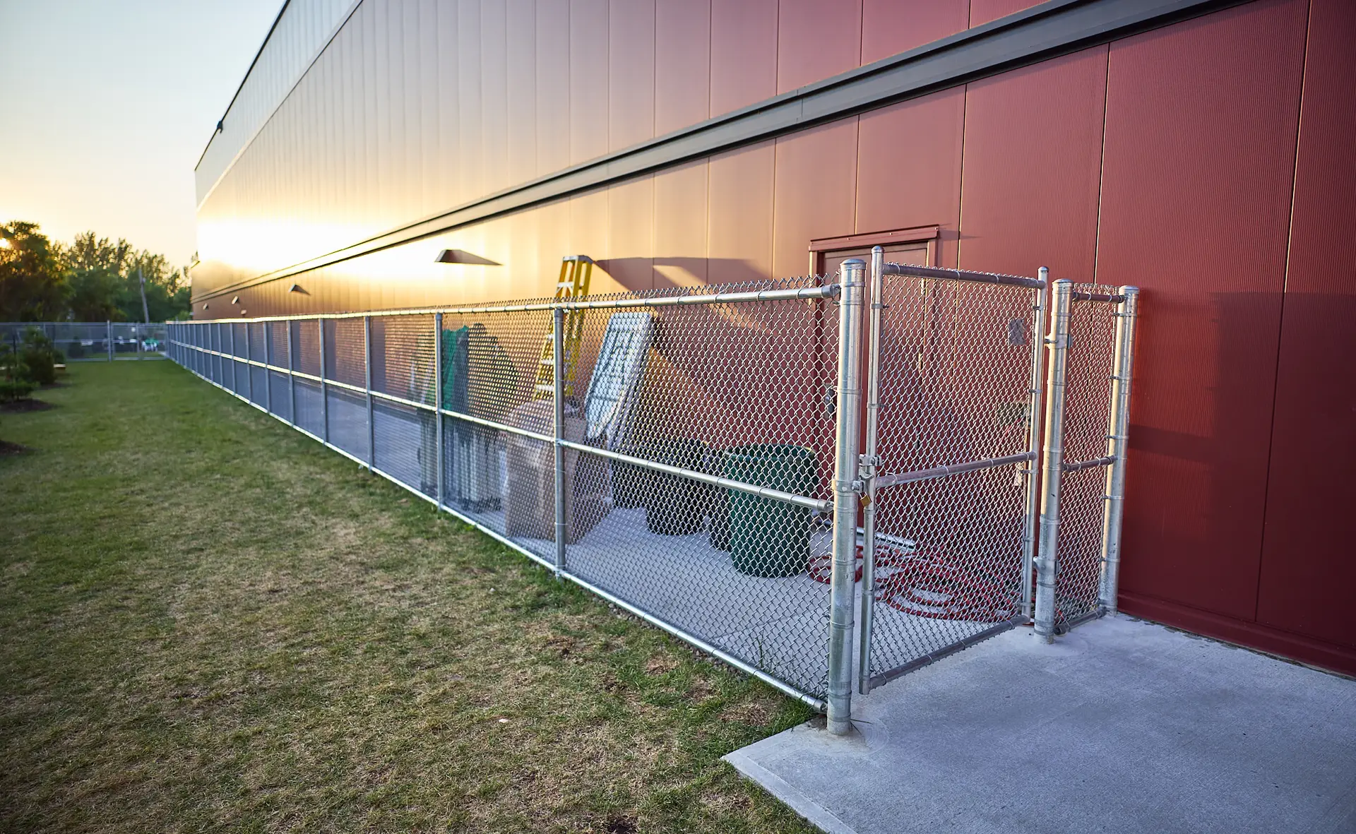 Inter Clôtures Sentinelle A chain-link fenced area beside a red industrial building contains trash bins, a ladder, and equipment. Fence Installation Quebec