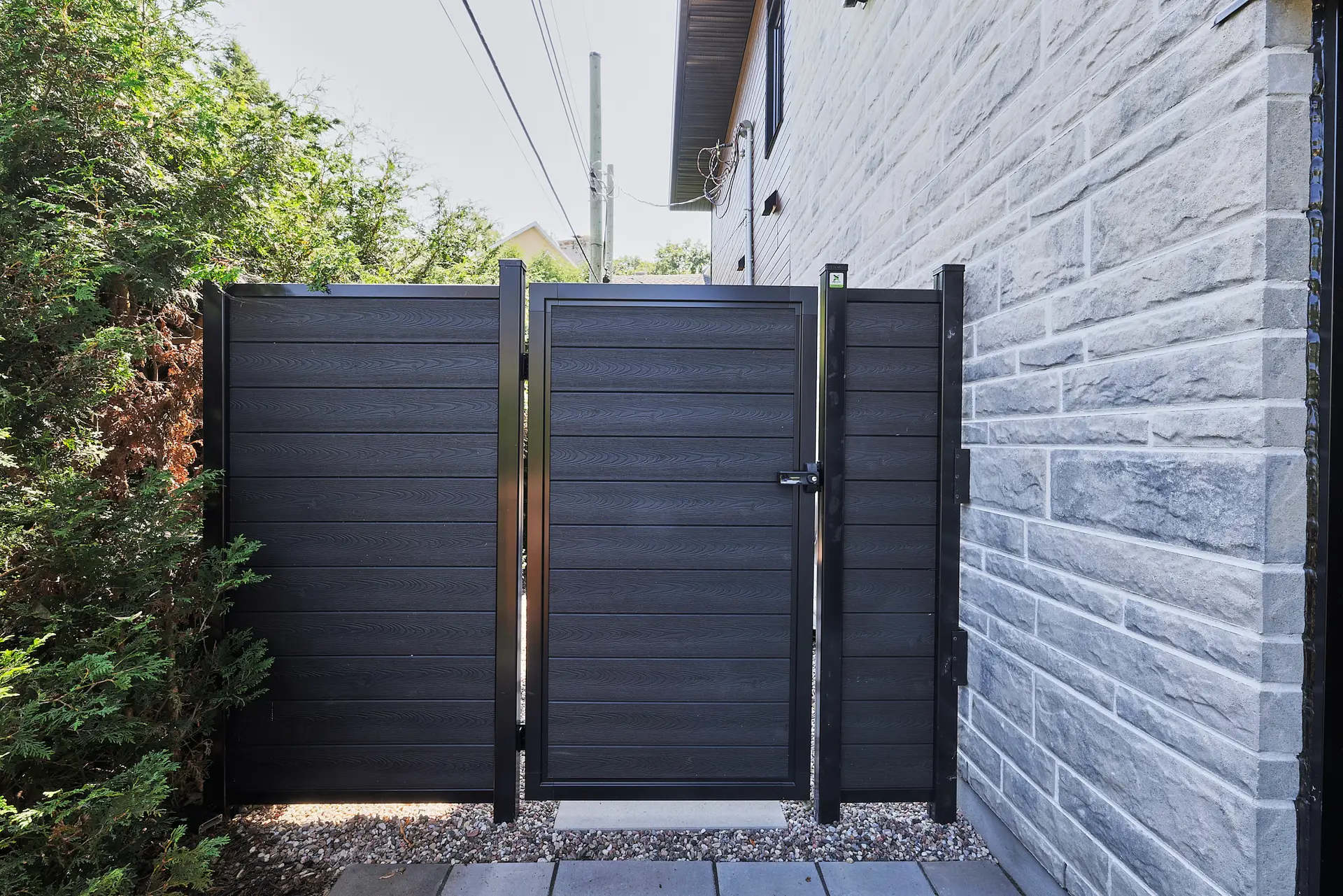 Inter Clôtures Sentinelle A black metal and composite fence gate stands between a stone building and green shrubs, with paving stones and gravel at the base. Fence Installation Quebec