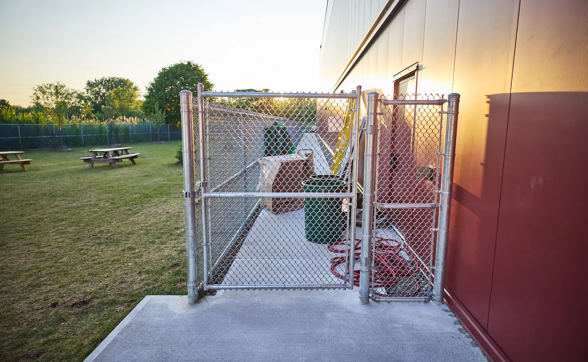 Inter Clôtures Sentinelle A fenced outdoor storage area next to a building, containing trash bins, hoses, and other equipment, with a grassy field and picnic tables in the background at sunset. Fence Installation Quebec