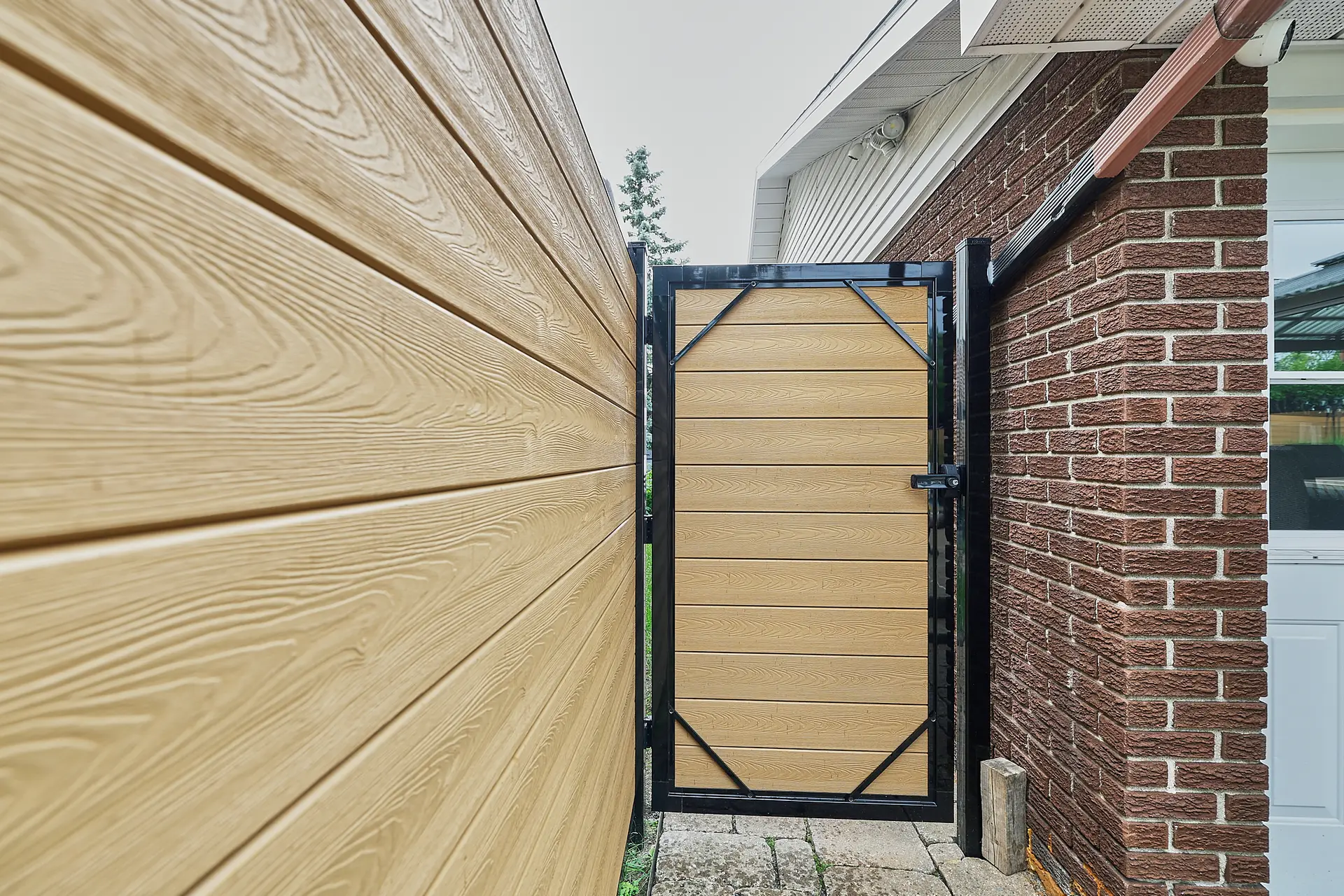 Inter Clôtures Sentinelle A wooden gate with black metal framing separates a brick house from a wooden fence; stone pavers cover the ground below. Fence Installation Quebec