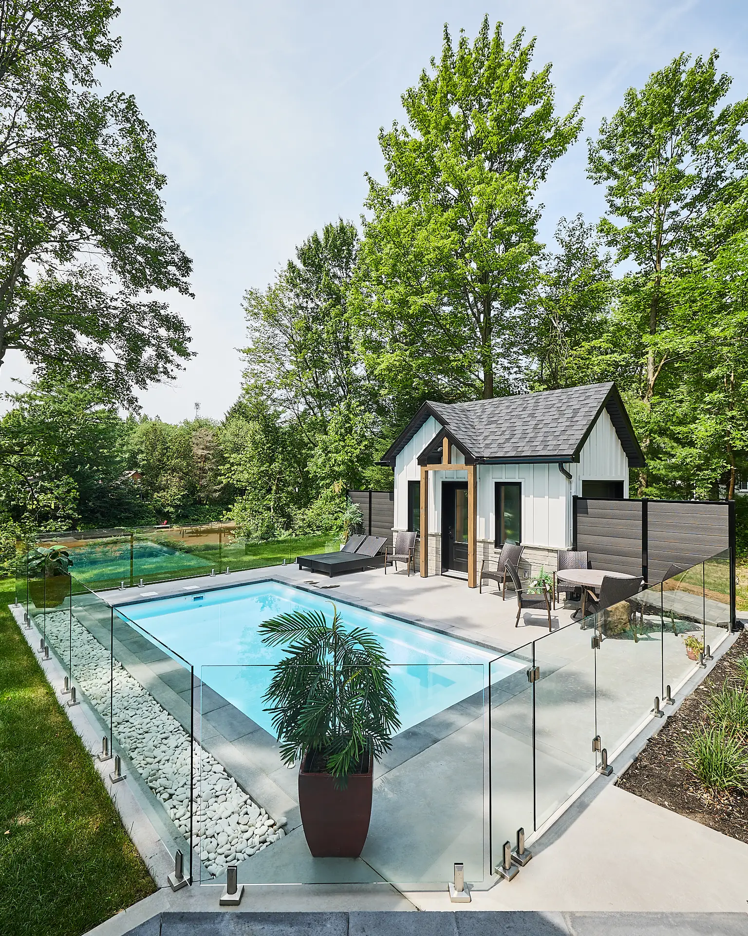 Inter Clôtures Sentinelle Modern pool area with glass fence, lounge chairs, potted plant, and a small pool house surrounded by trees and greenery on a sunny day. Fence Installation Quebec