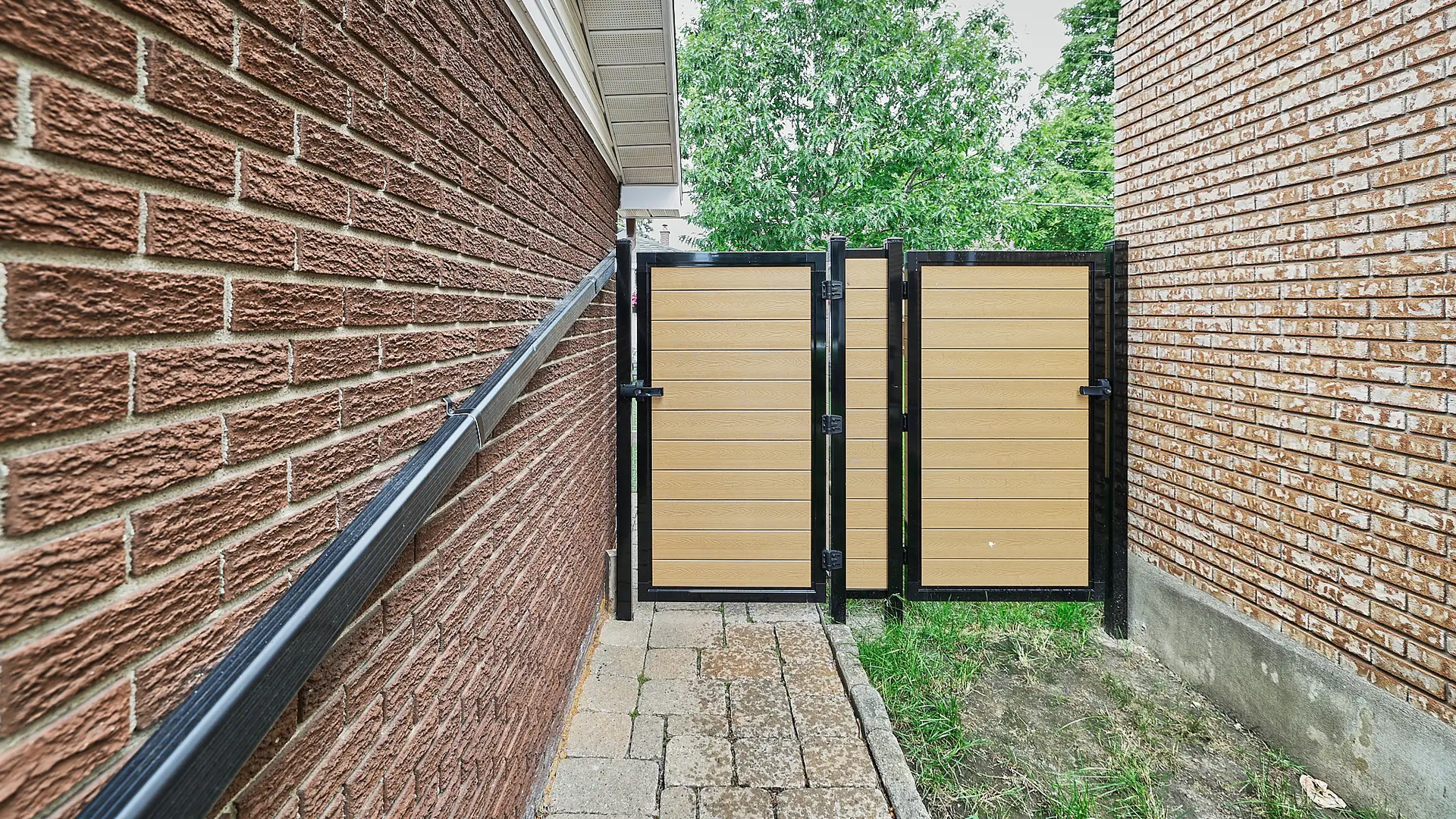 Inter Clôtures Sentinelle A black metal and wood gate stands between two brick buildings on a stone pathway with grass to one side. Fence Installation Quebec