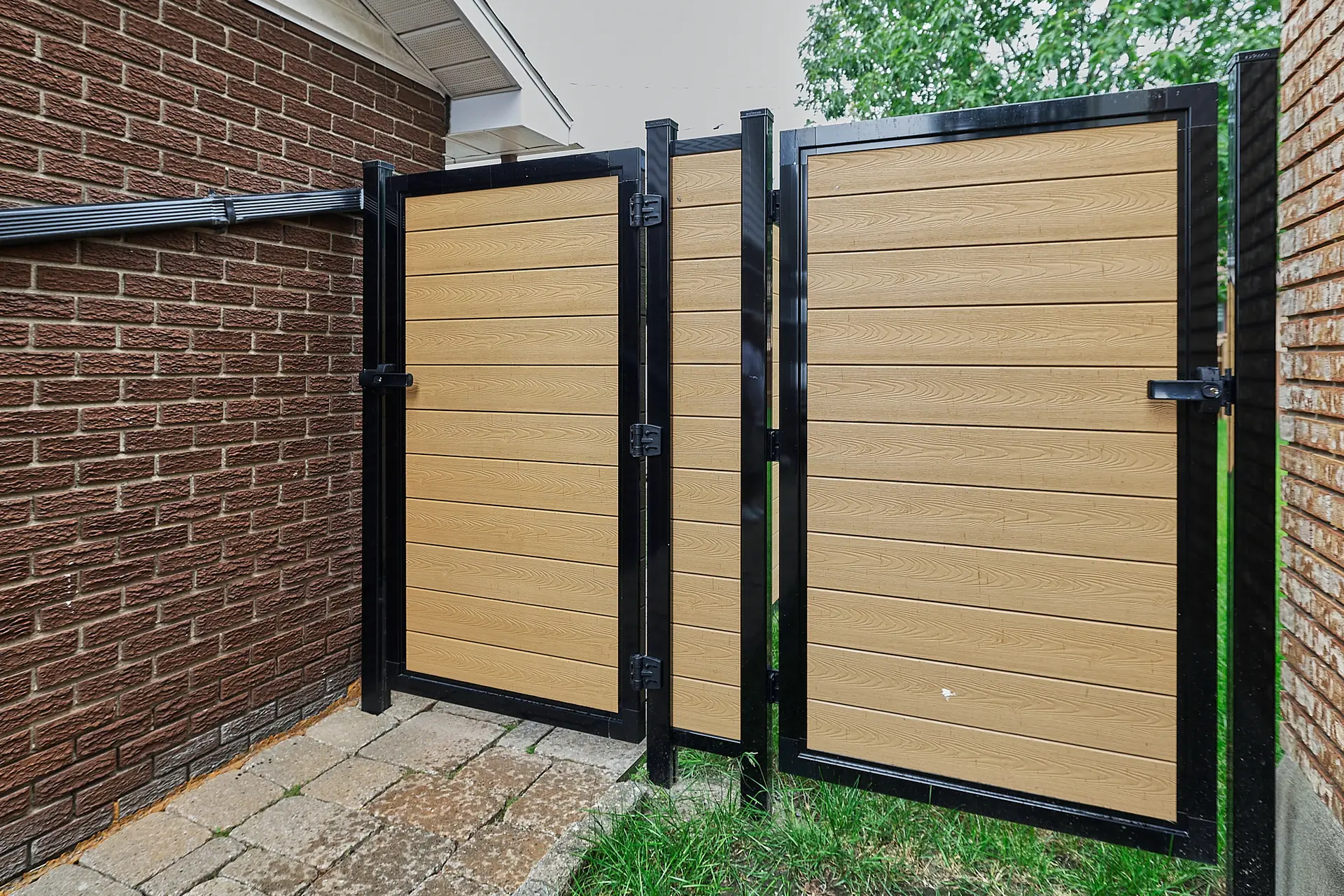 Inter Clôtures Sentinelle A modern gate with beige horizontal panels and a black frame stands between two brick walls beside a paved pathway and grass. Fence Installation Quebec