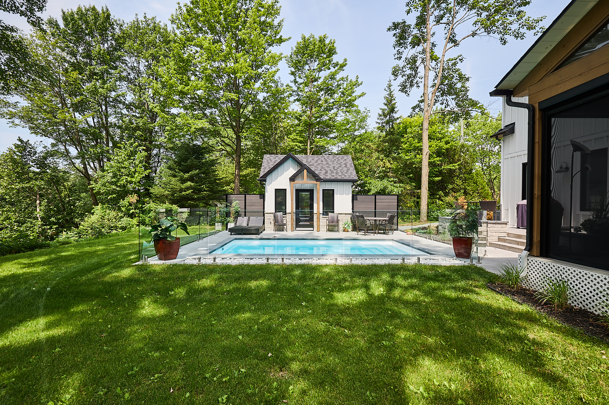 Inter Clôtures Sentinelle A small rectangular pool in a backyard with glass fencing, lounge chairs, potted plants, and a pool house, surrounded by trees and grass. Fence Installation Quebec