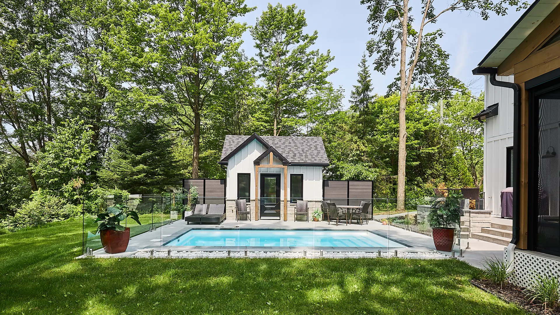 Inter Clôtures Sentinelle A small in-ground pool with clear fencing, surrounded by grass, sits in a backyard with trees and a modern pool house in the background. Two lounge chairs and large potted plants are nearby. Fence Installation Quebec