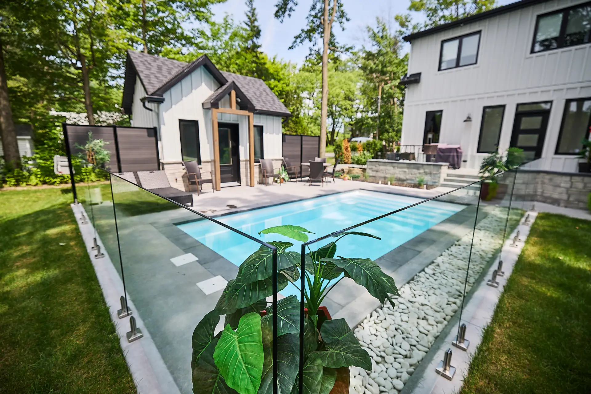 Inter Clôtures Sentinelle A small backyard pool is surrounded by a glass fence, with a modern guesthouse and main house in the background. Large potted plants and patio furniture are also visible. Fence Installation Quebec
