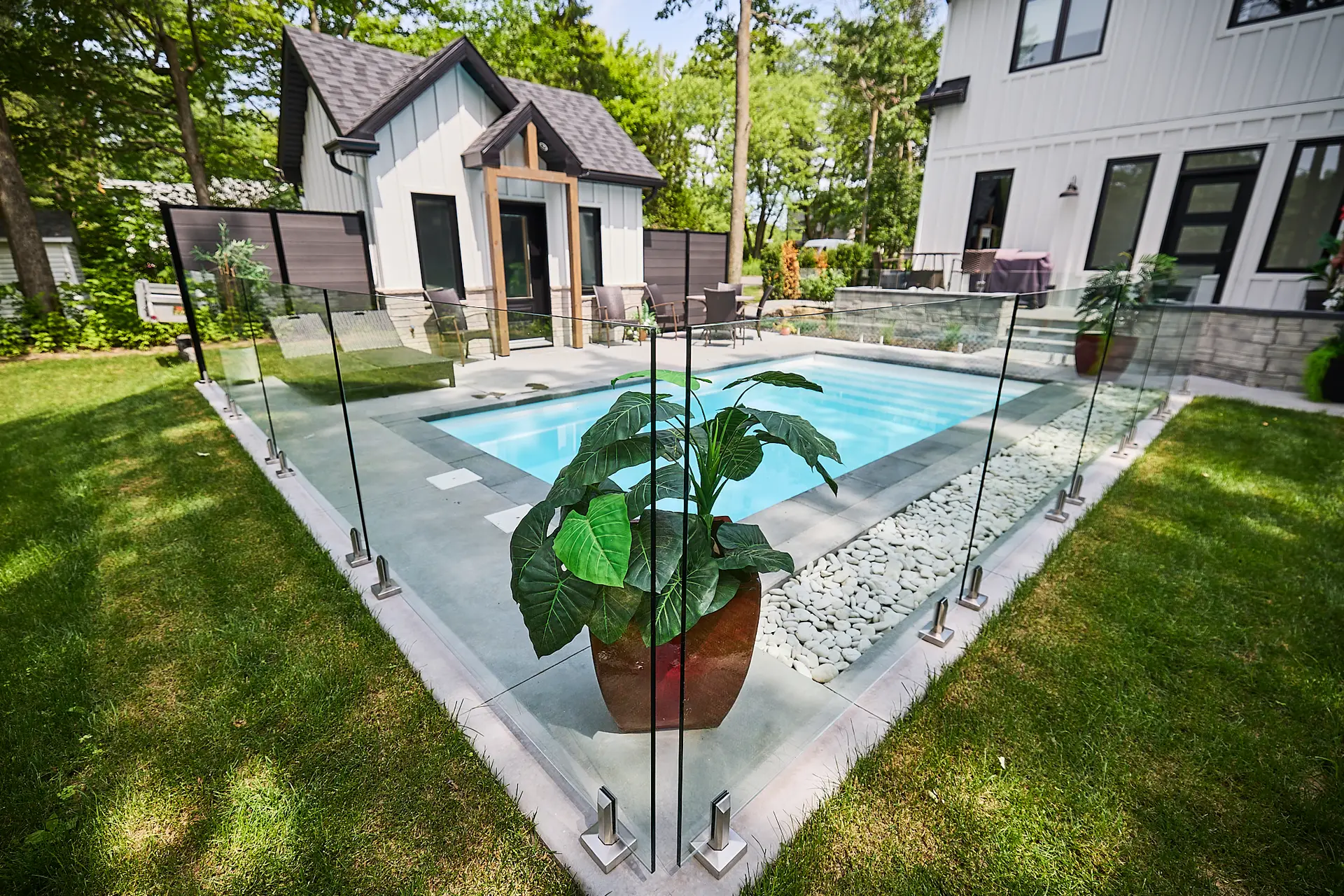 Inter Clôtures Sentinelle Small backyard pool surrounded by glass fencing, with a potted plant in the foreground, green lawn, and two modern buildings in the background. Fence Installation Quebec