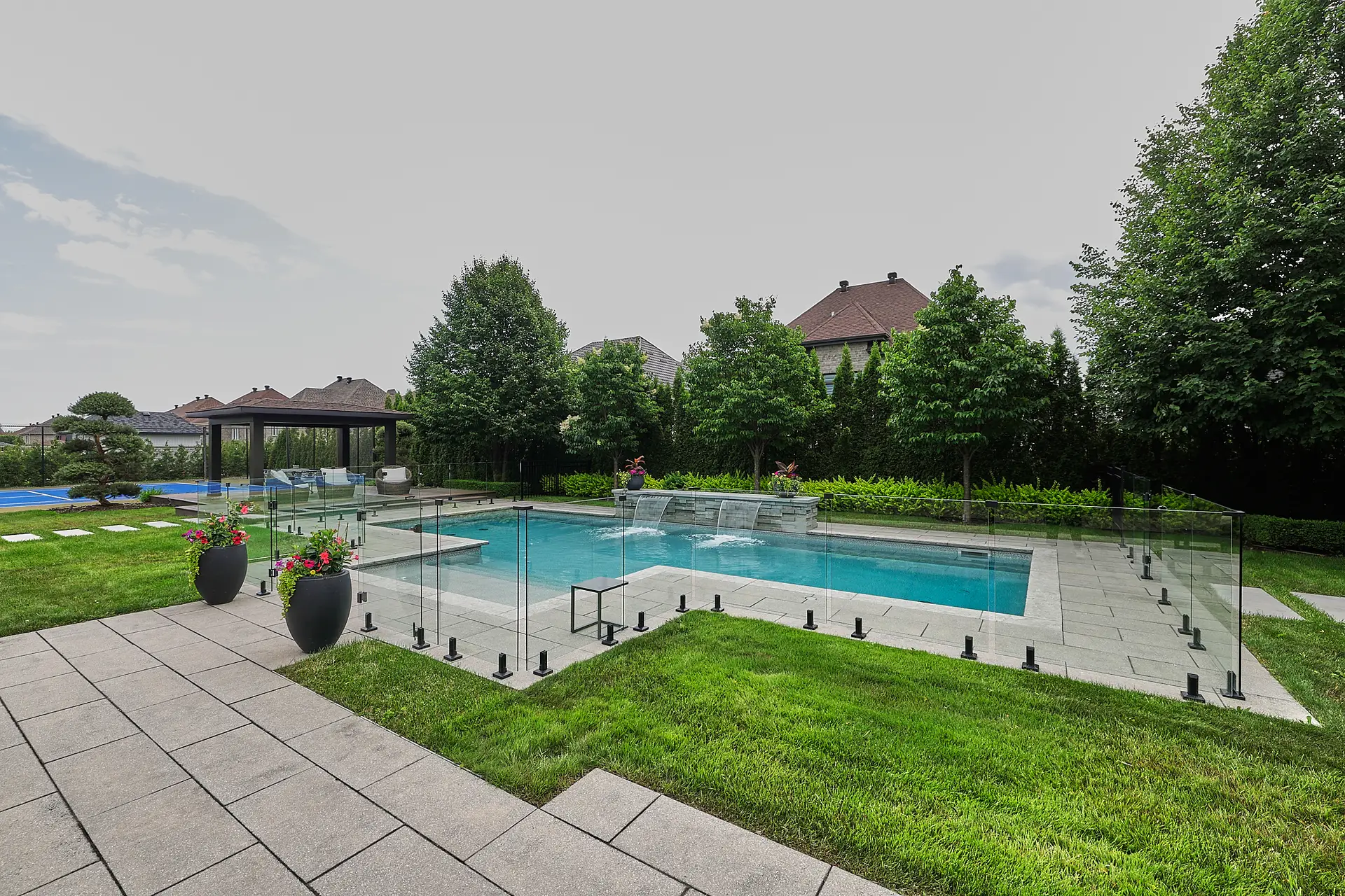 Inter Clôtures Sentinelle Rectangular outdoor pool with glass fencing, surrounded by grass and pavers, with trees and houses in the background under a cloudy sky. Fence Installation Quebec