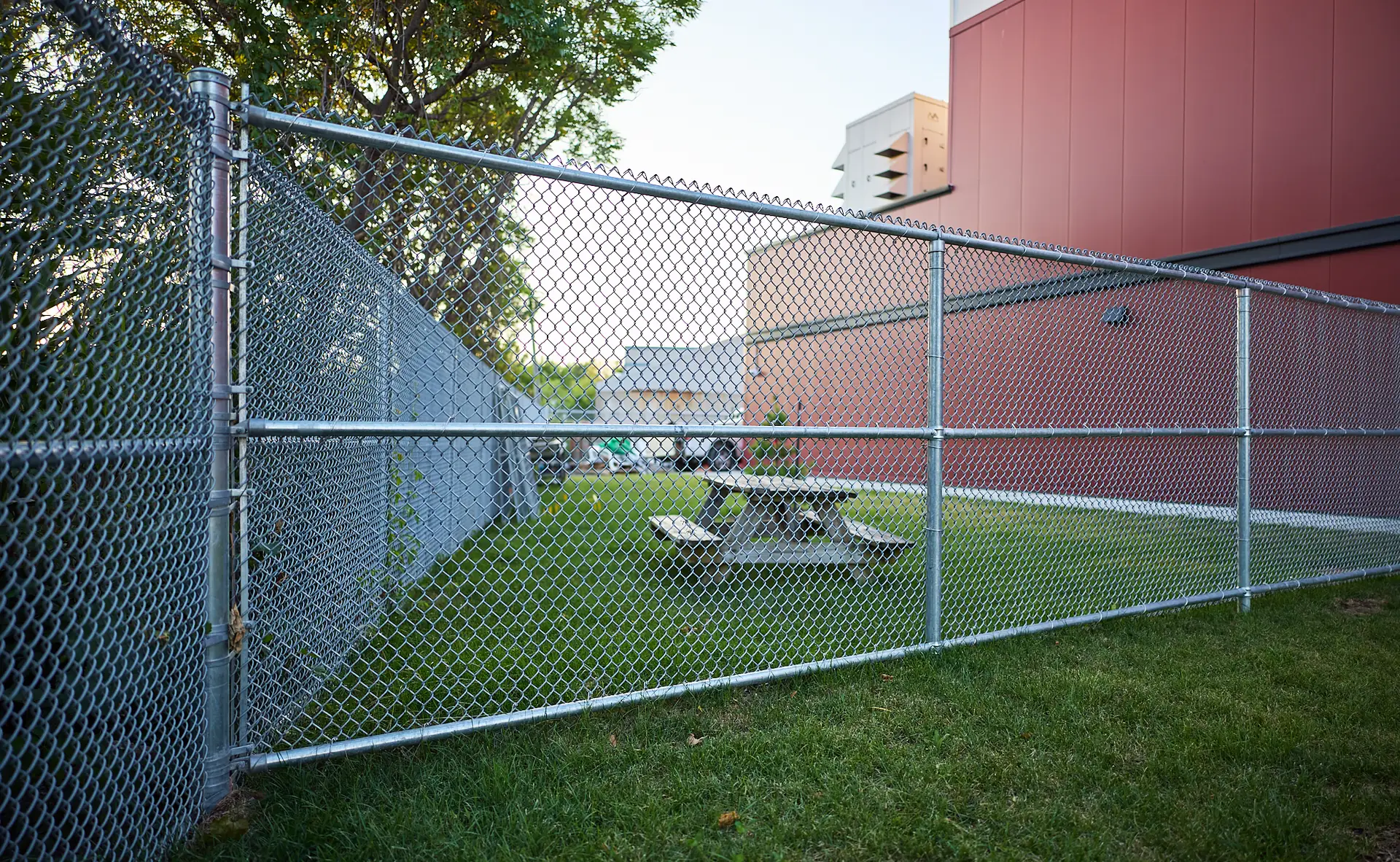 Inter Clôtures Sentinelle A fenced grassy area with a picnic table, next to a red building and trees, on a clear day. Fence Installation Quebec