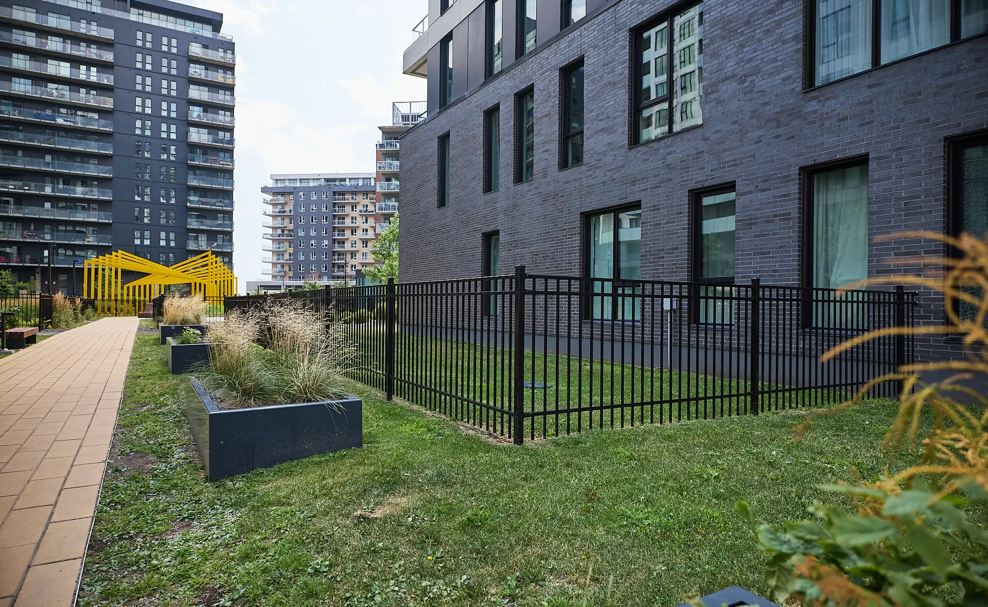 Inter Clôtures Sentinelle Modern apartment buildings with large windows and balconies, a fenced grassy area, potted plants, and a yellow structure on a paved walkway in an urban setting. Fence Installation Quebec