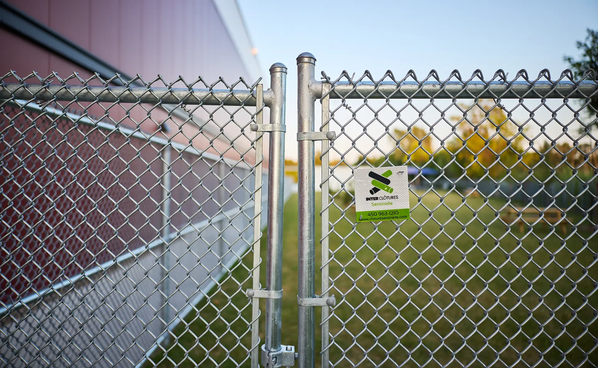 Inter Clôtures Sentinelle A closed chain-link gate with a lock and a green sign is in front of a grass area next to a red building. Fence Installation Quebec