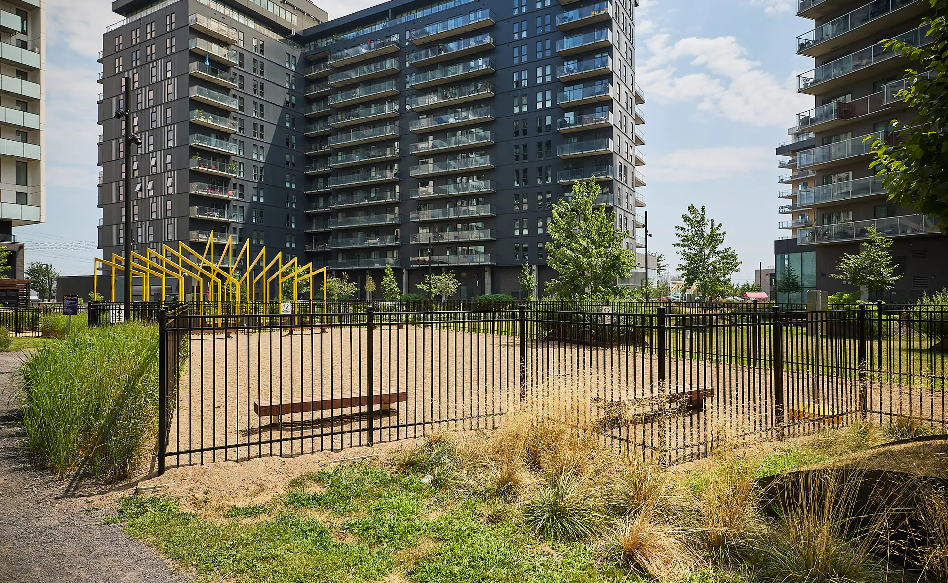 Inter Clôtures Sentinelle A fenced sandy dog park with benches is surrounded by tall modern apartment buildings, greenery, and a yellow metal structure in the background. Fence Installation Quebec