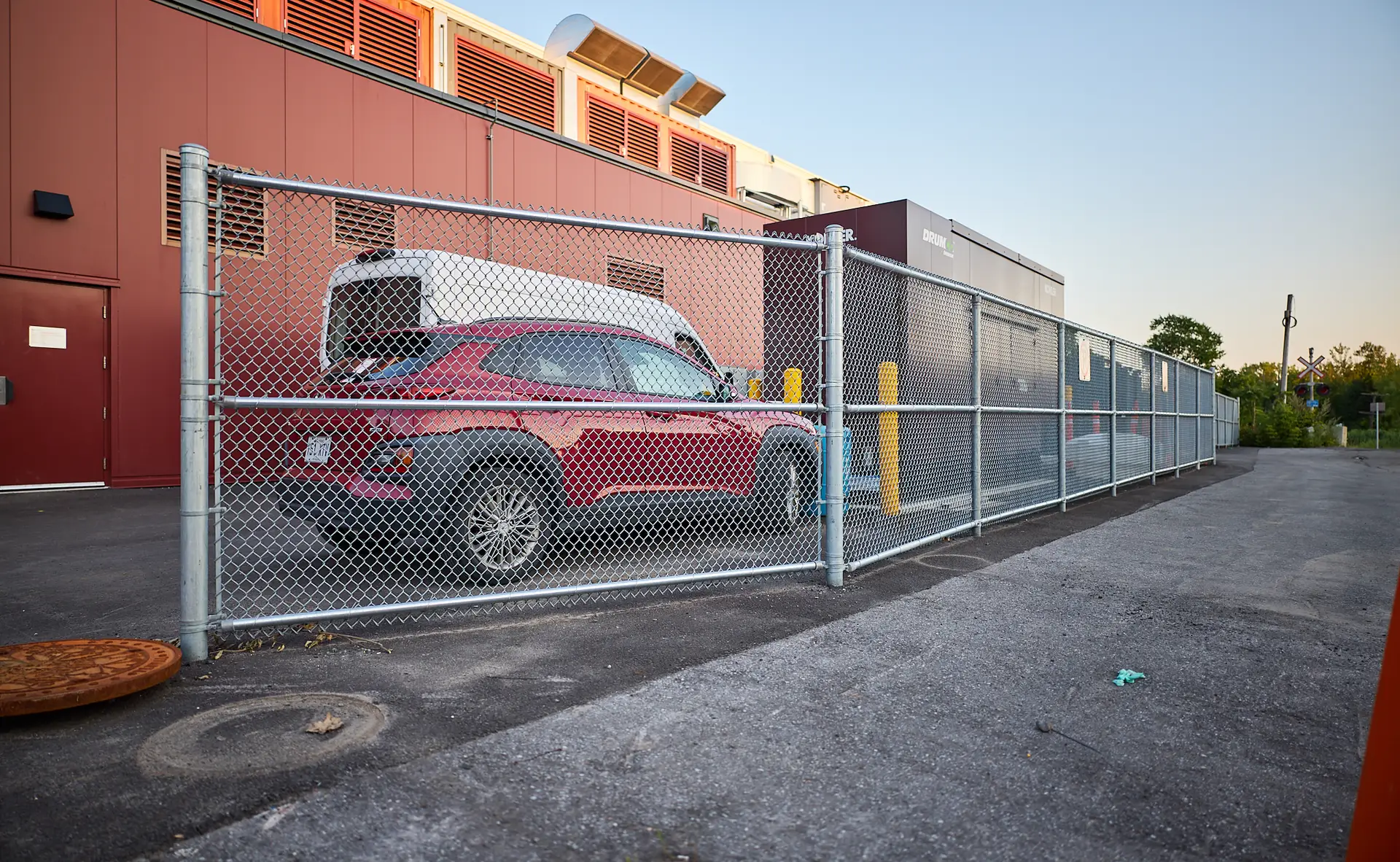 Inter Clôtures Sentinelle A red and white vehicle is parked behind a chain-link fence next to a red industrial building with vents and doors. Fence Installation Quebec