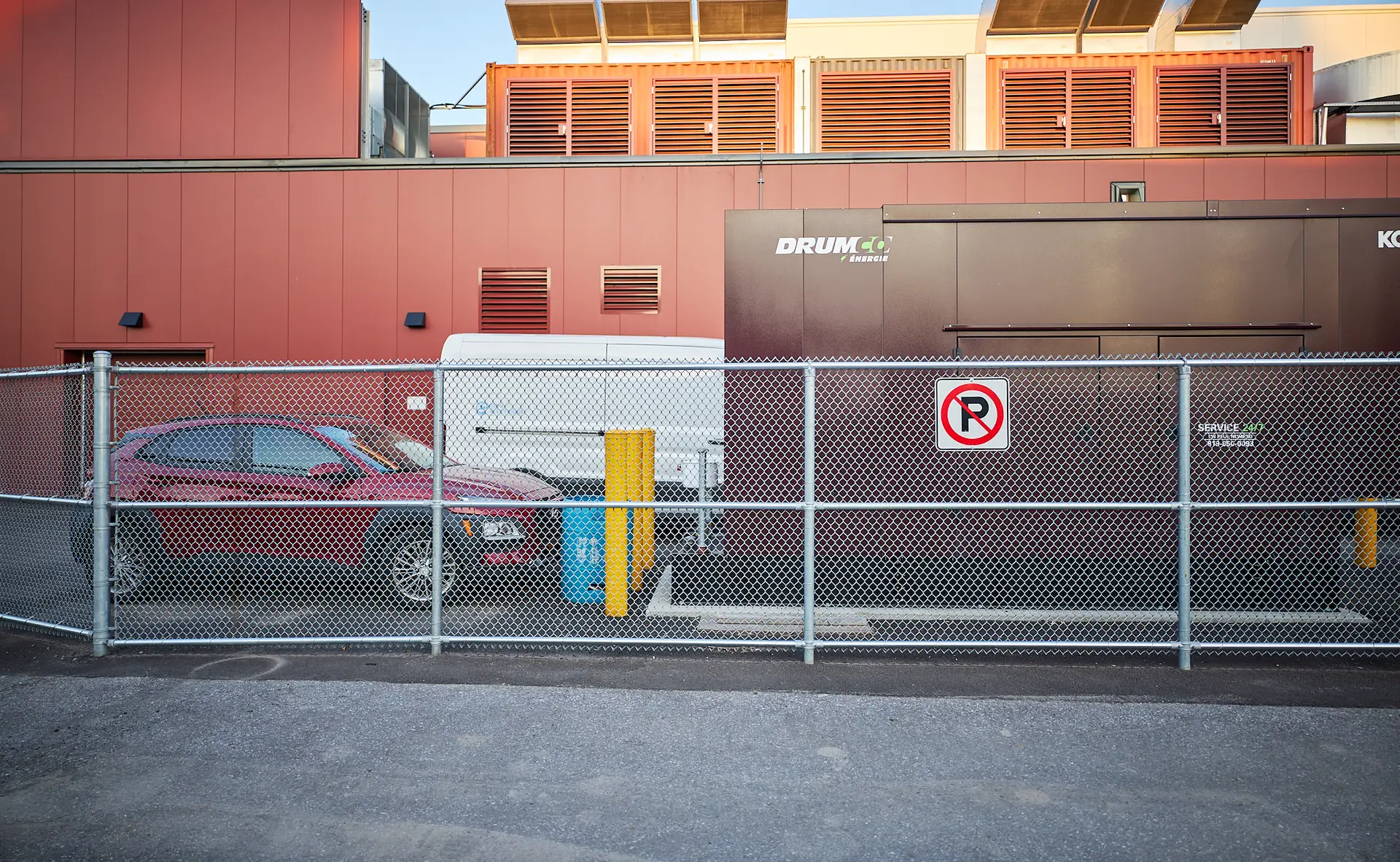 Inter Clôtures Sentinelle A red car and white van are parked behind a chain-link fence next to a building, with a “No Parking” sign visible on a dark container. Fence Installation Quebec