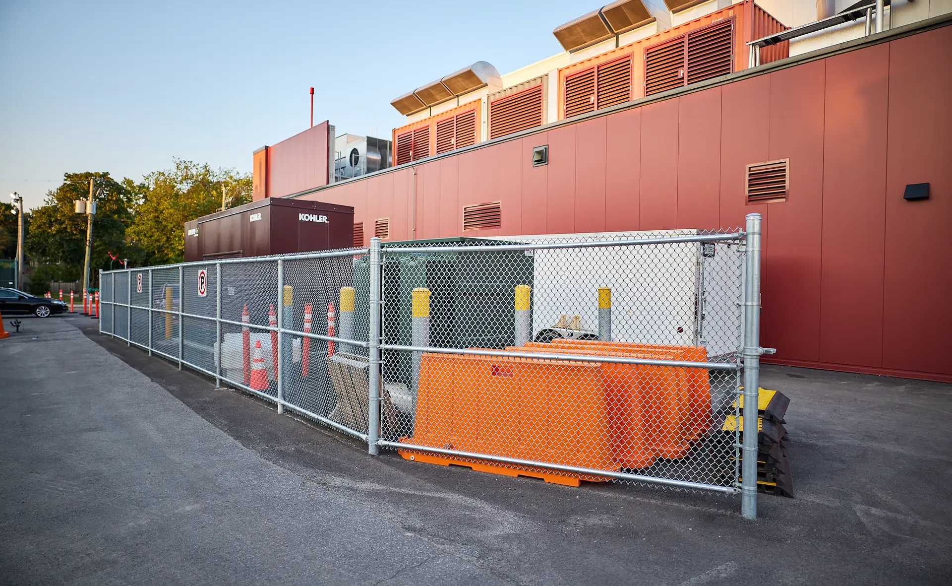 Inter Clôtures Sentinelle A fenced-in utility area with orange barriers and traffic cones next to a red industrial building; no entry signs are visible. Fence Installation Quebec