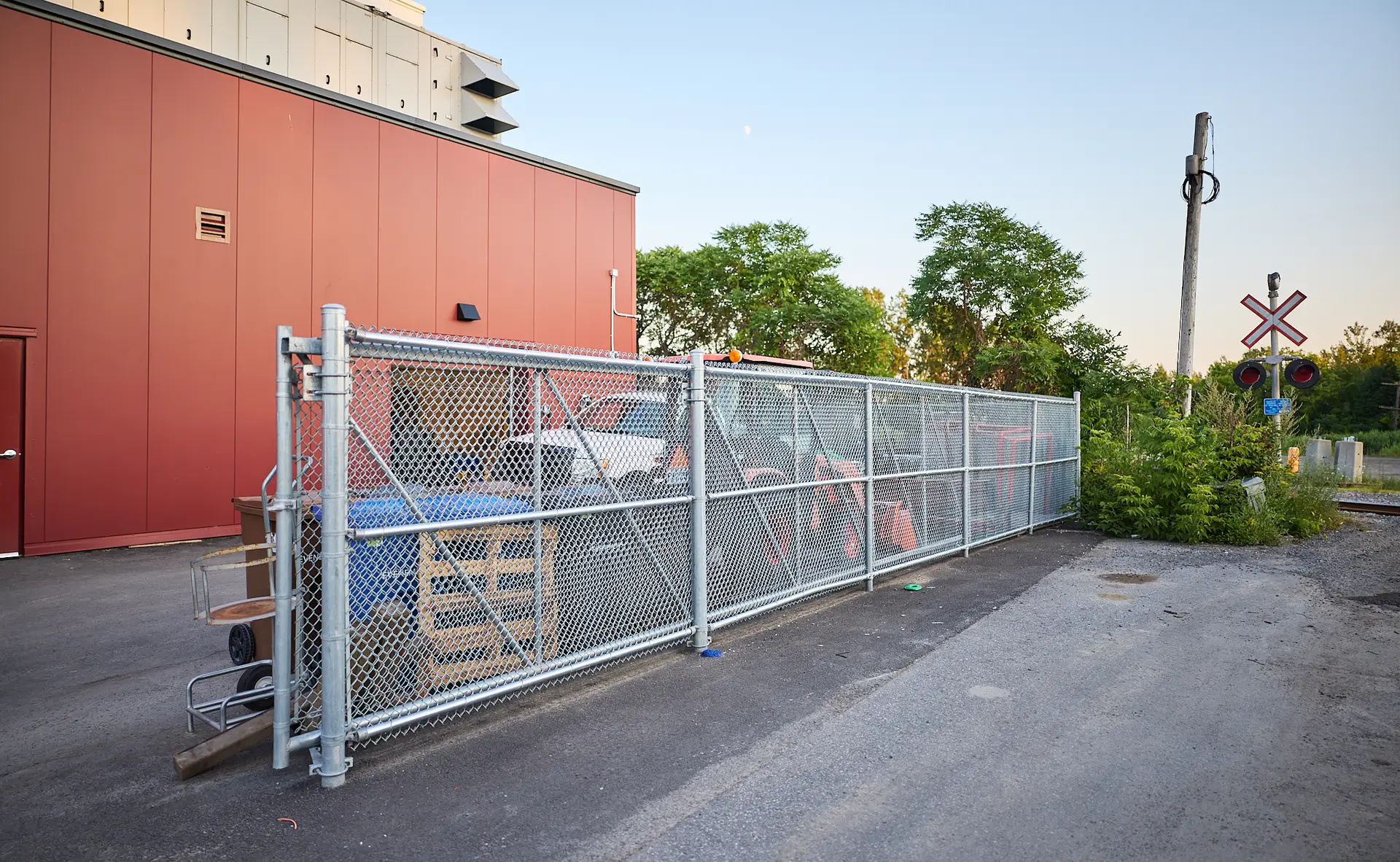Inter Clôtures Sentinelle A fenced enclosure beside a red building contains various items and is located near a railroad crossing with visible signal posts and greenery in the background. Fence Installation Quebec