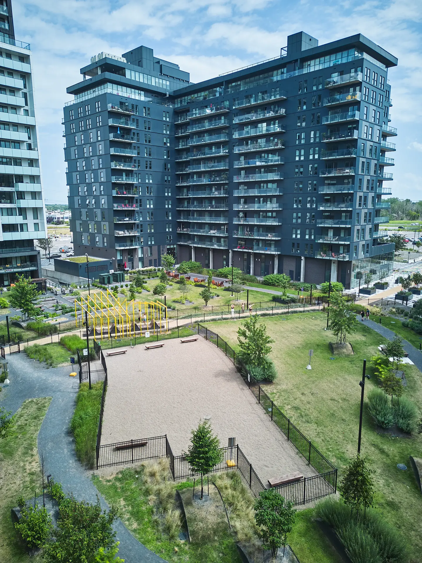 Inter Clôtures Sentinelle A modern apartment complex overlooks a landscaped courtyard with a fenced sand area, playground equipment, walking paths, and scattered trees on a clear day. Fence Installation Quebec