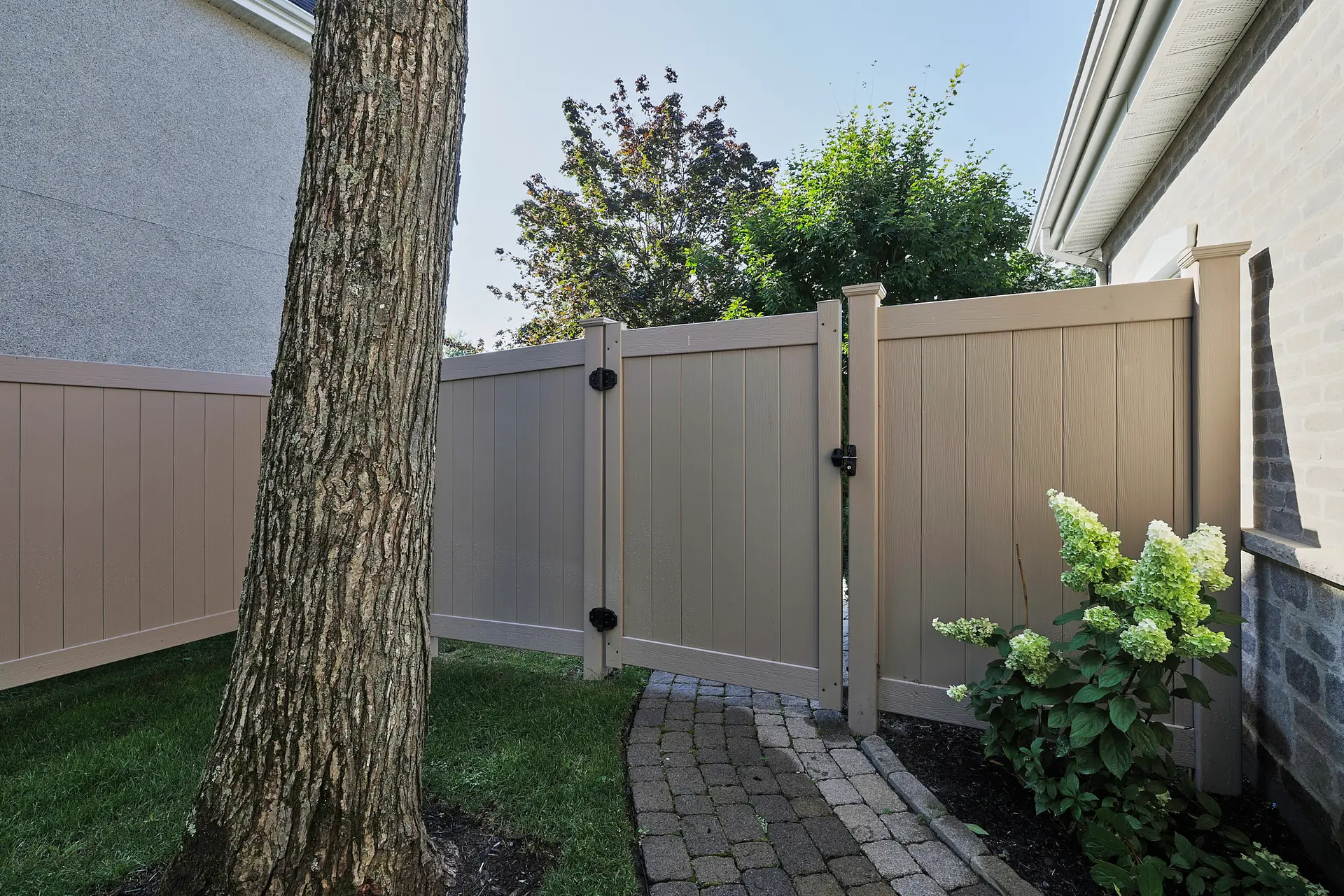 Inter Clôtures Sentinelle A beige wooden fence with a closed gate encloses a small backyard, featuring a tree, green grass, a hydrangea plant, and a curved stone pathway. Fence Installation Quebec