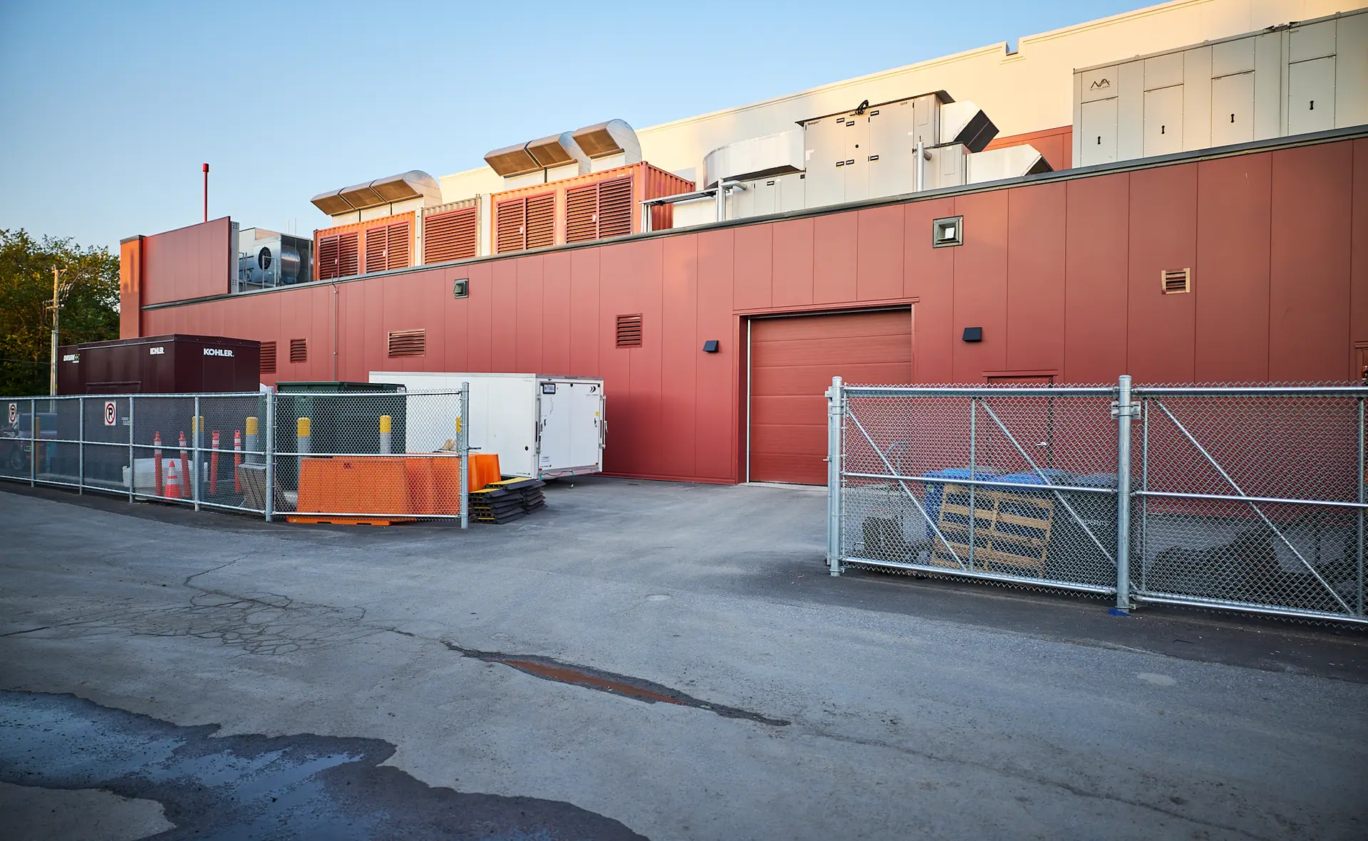 Inter Clôtures Sentinelle A fenced utility area behind a red industrial building with HVAC units on the roof, parked equipment, and a closed garage door. Fence Installation Quebec