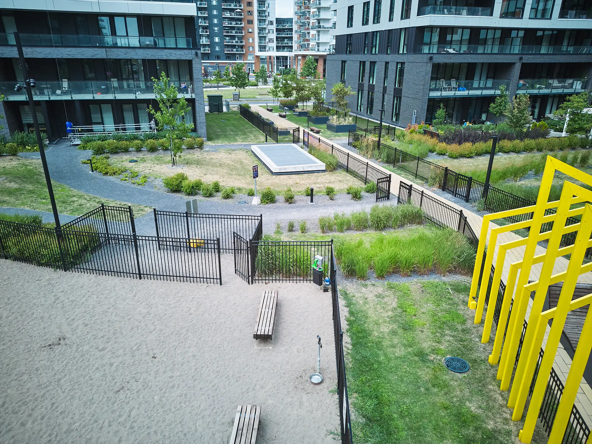 Inter Clôtures Sentinelle A fenced urban park area with benches, sand, a path, grassy sections, yellow structures, and modern apartment buildings in the background. Fence Installation Quebec