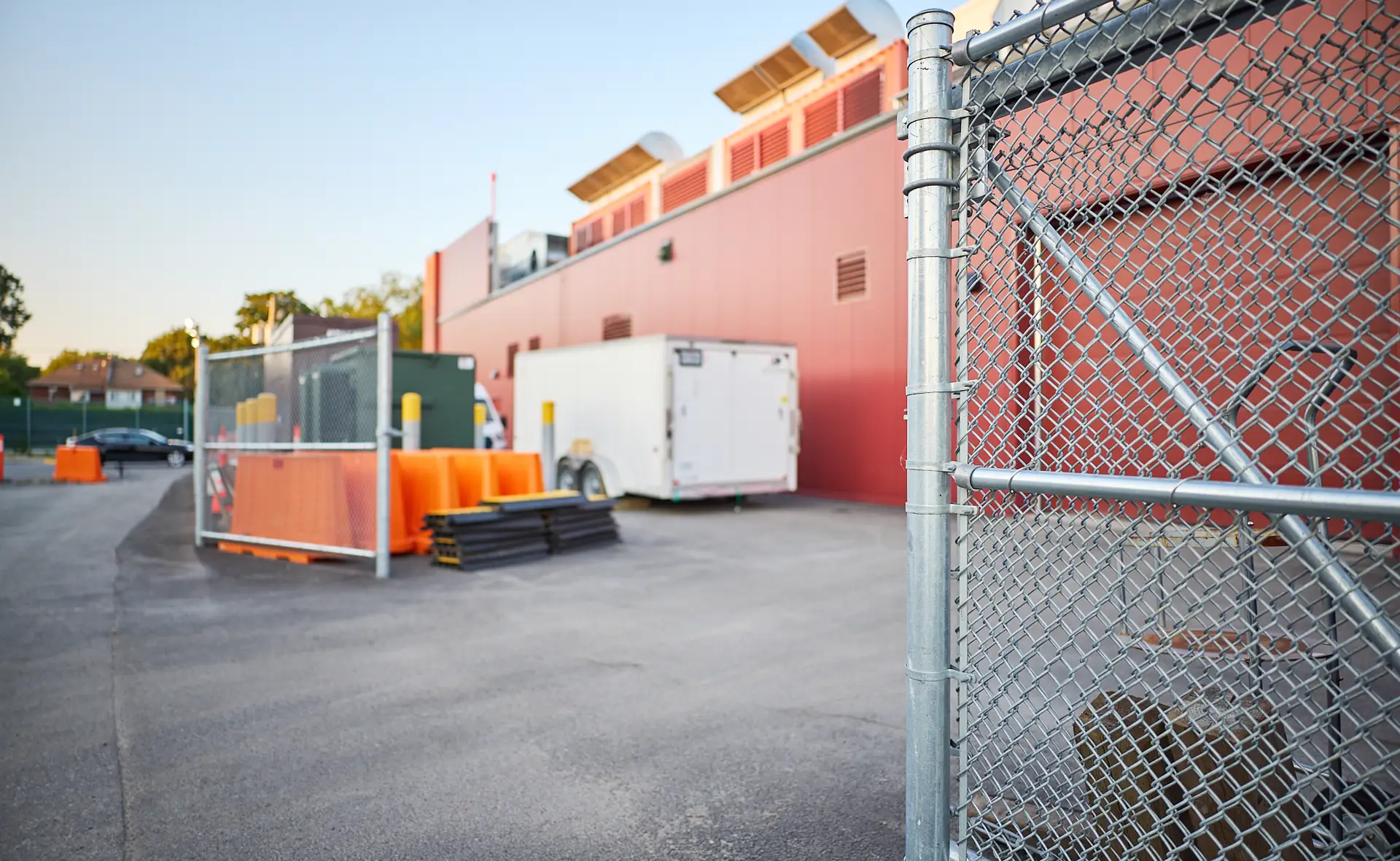 Inter Clôtures Sentinelle A fenced-off area next to a red industrial building with an equipment trailer, orange barriers, and metal fencing on an asphalt surface. Fence Installation Quebec