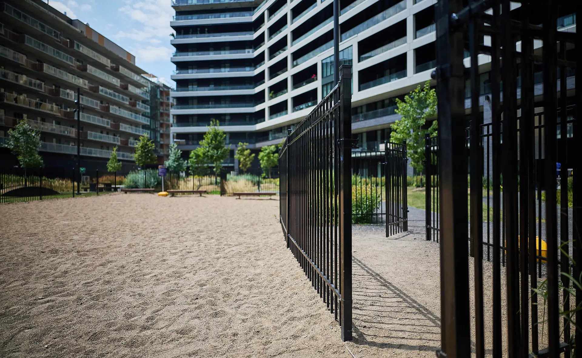 Inter Clôtures Sentinelle A gated sandy area with trees is surrounded by modern apartment buildings on a sunny day. Fence Installation Quebec
